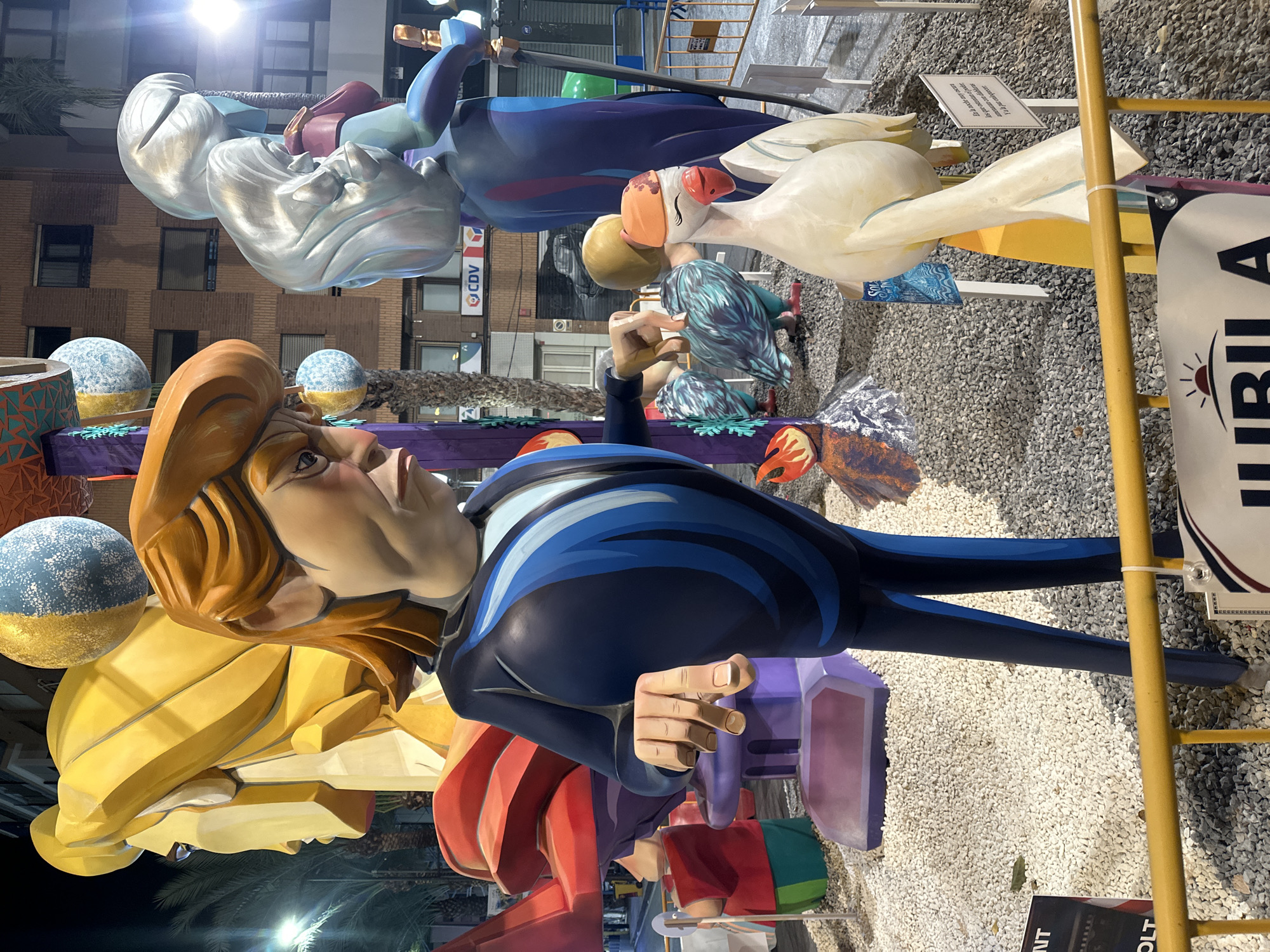 This image depicts an outdoor nighttime scene at a festival in Alicante, Spain. The foreground features several large, colorful sculptures. A prominent figure, appearing to be a caricature of Donald Trump, is center-left, wearing a dark blue suit with blue accents. He is gesturing with his right hand, fingers extended. To his right is a white goose sculpture wearing an orange hat and with its eyes closed. In the background, other sculptures are visible, including a large yellow head on the left, a silver-haired figure on the right, and two stylized birds in blue and green. The ground is covered with gravel. A yellow barricade runs across the bottom of the image. Buildings and streetlights are visible in the background. The overall impression is a festive and somewhat surreal display.