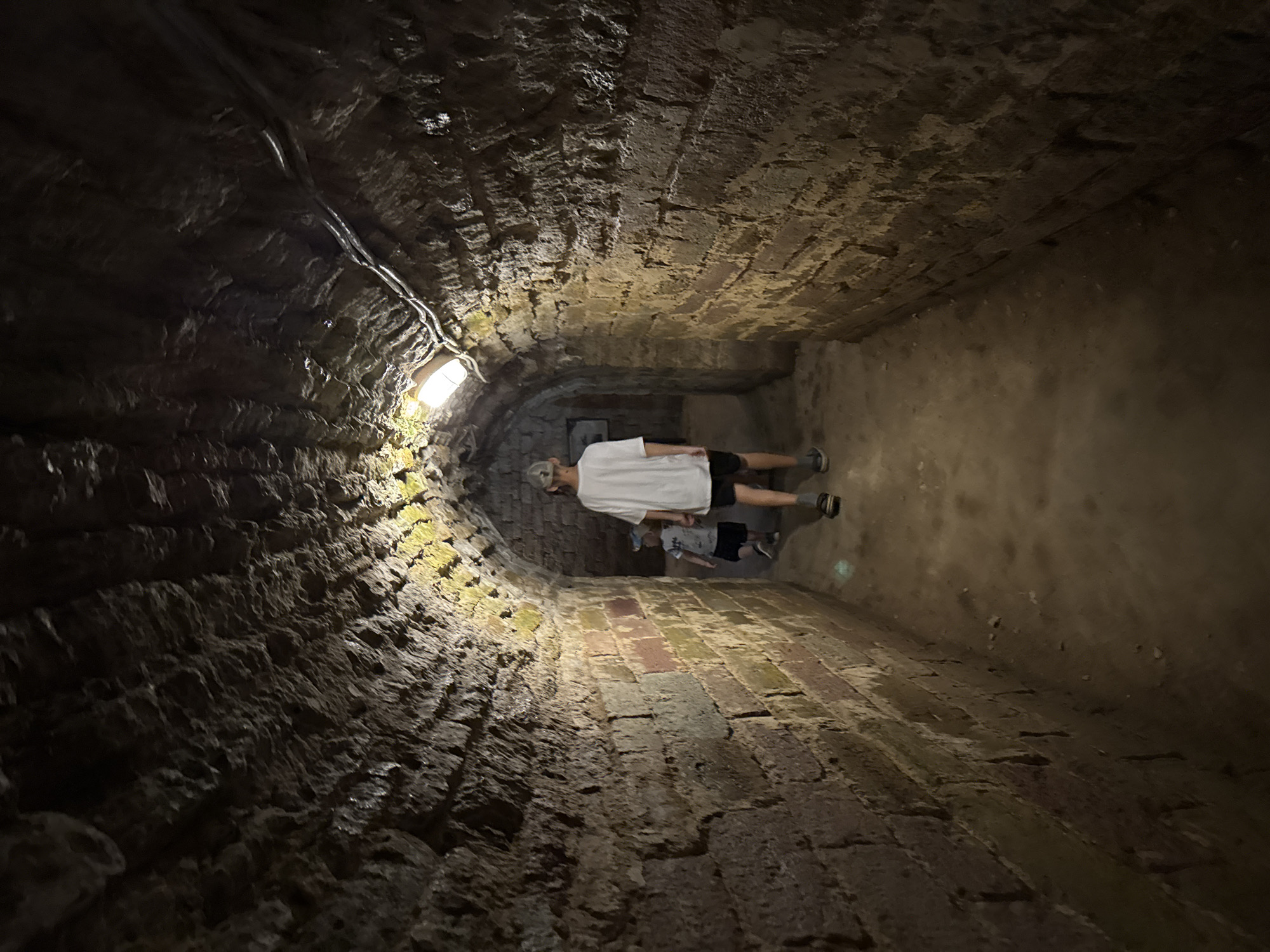 Two individuals, an adult and a child, are walking through a dimly lit, arched stone tunnel. The tunnel walls are constructed from rough-hewn, large stone blocks, some of which appear to be reddish-brown and others a lighter, sandy color, with patches of moss or algae. The ceiling of the tunnel is also made of stone and appears roughly shaped. Two modern, rectangular light fixtures are visible on the ceiling, providing illumination. The floor of the tunnel is dirt or gravel. The adult, wearing a white t-shirt, black shorts, and a light-colored baseball cap, is walking ahead of the child, who is wearing a dark t-shirt, shorts, and a blue baseball cap. The image was taken in Posyolok Imeni Morozova, Russia.