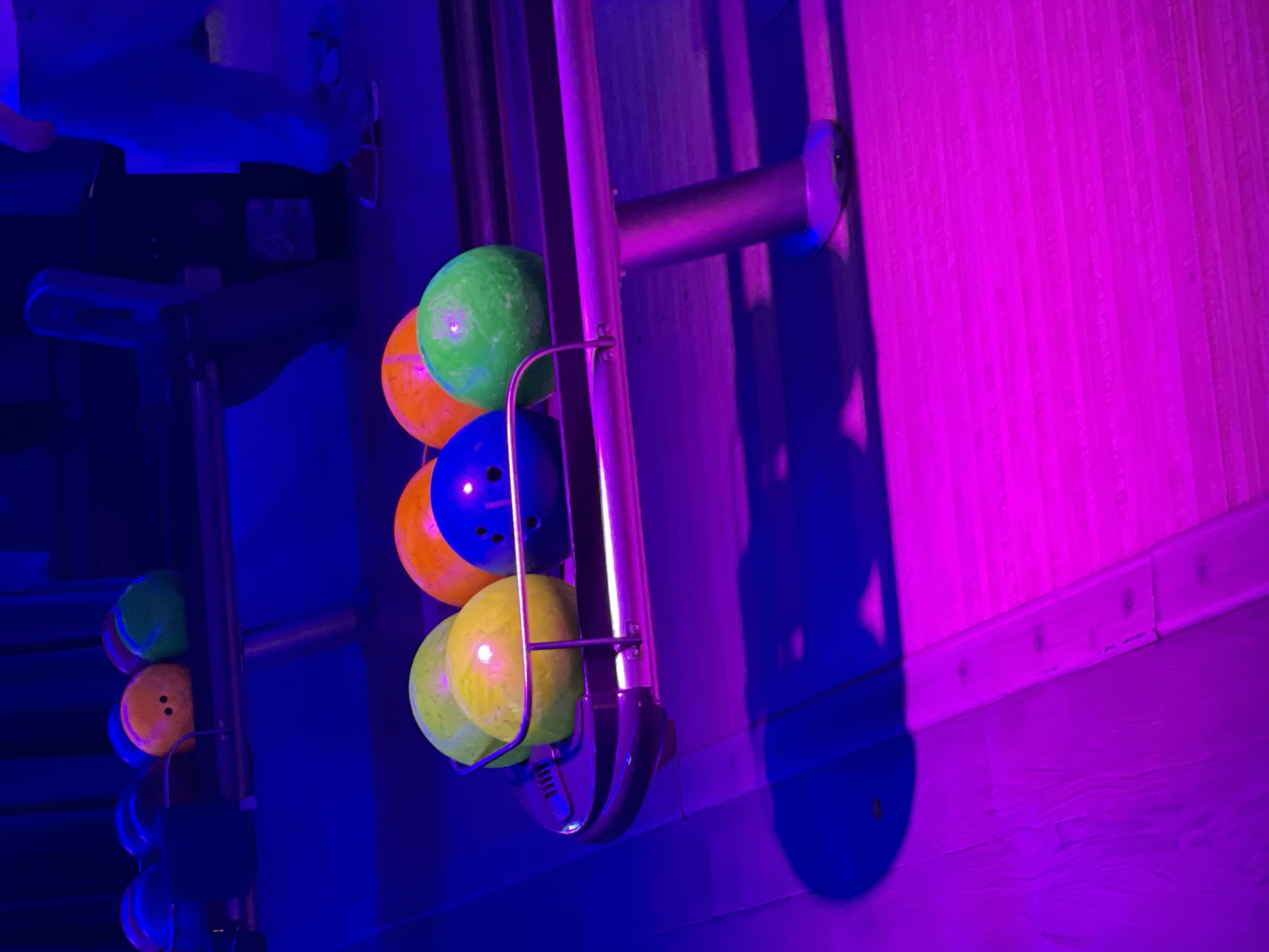 The image shows bowling balls on a rack in what appears to be a bowling alley. There are different colored bowling balls, including green, orange, blue, and yellow. The bowling ball rack is a silver metal. The lighting is blue and pink. Based on the location provided in the prompt, the image is taken in Sochi, Russia.