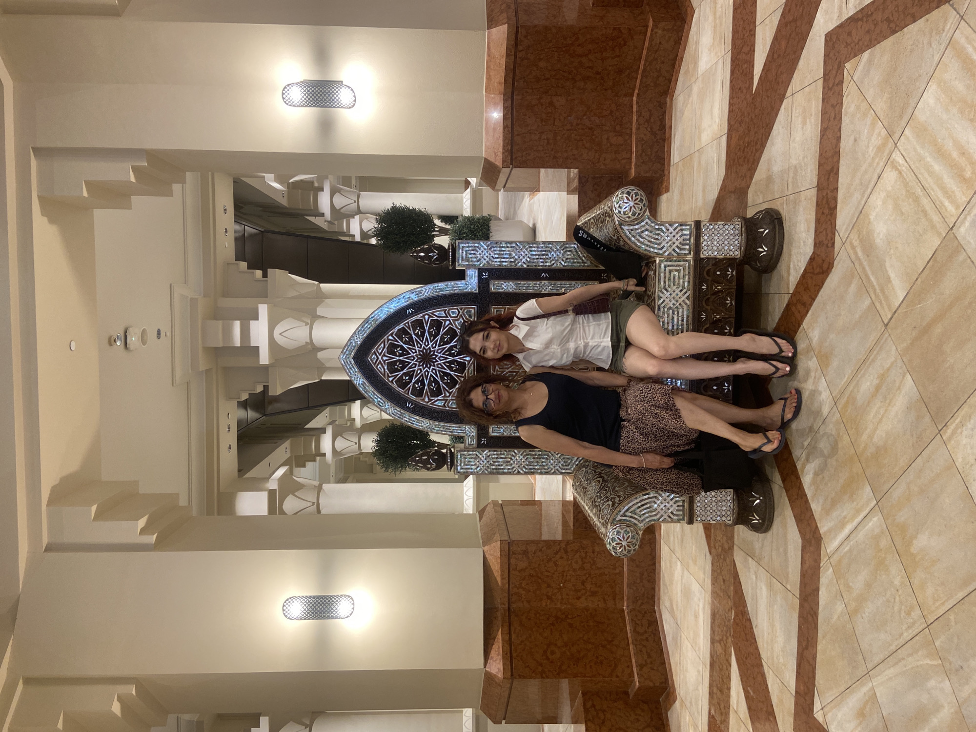 The image depicts two women posing on a decorative, mosaic-inlaid throne or bench. They are located within a large, ornate lobby or public space. The women, likely a mother and daughter, are dressed in casual clothing, one in a white top and olive shorts, the other in a black tank top and patterned skirt.
The architectural details suggest a location within a luxury hotel or resort in Dubai, United Arab Emirates. Architectural elements include geometric patterns, archways, and decorative lighting. The flooring features a geometric pattern. A decorative archway and staircases are visible in the background.