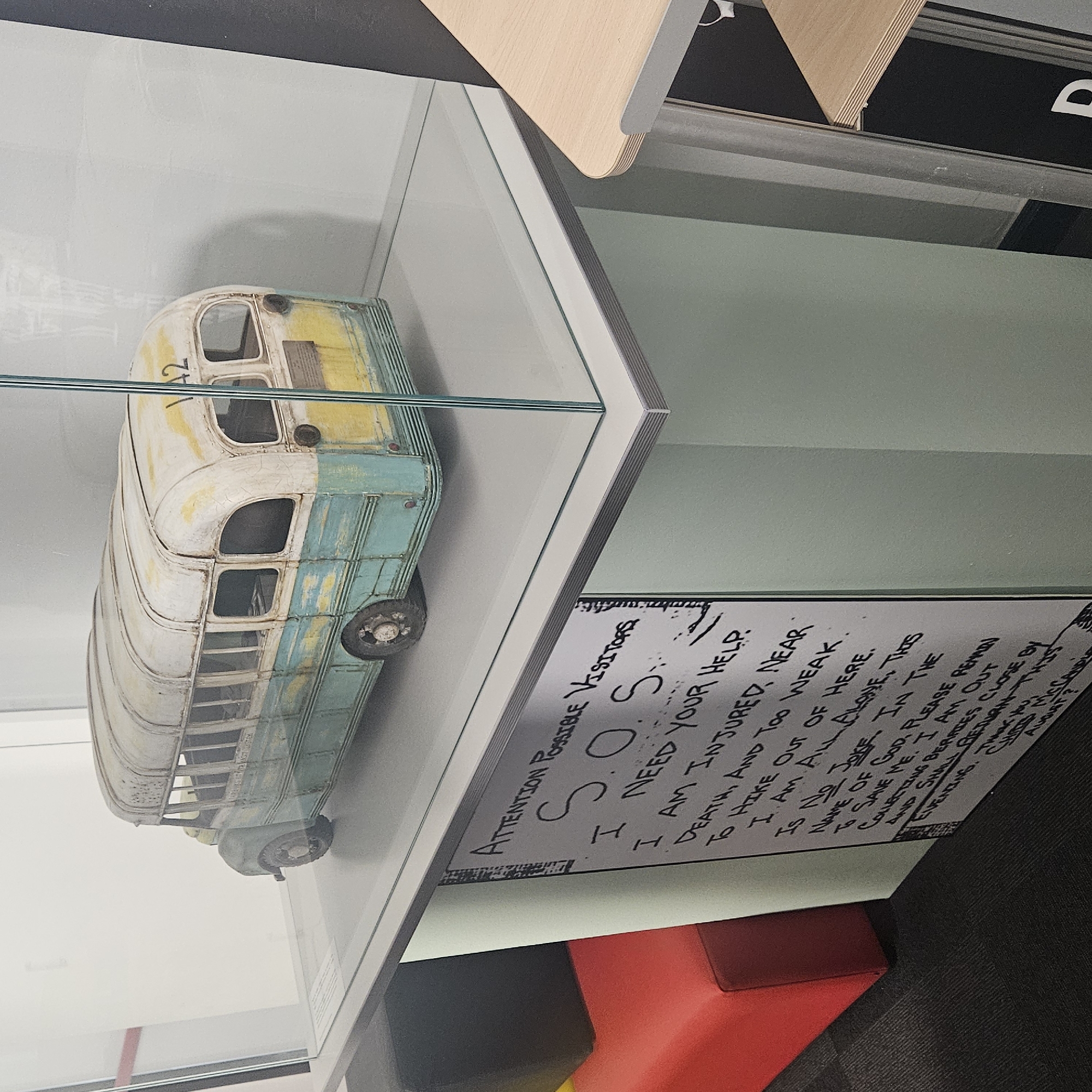 The image shows a museum display featuring a model of a vintage school bus. The bus is weathered, with peeling paint revealing a pale blue and yellow undercoat. It has the number "142" visible on its rear. The model is housed within a glass display case. Below the display case, a sign is affixed to a light green vertical panel. The sign, written in black marker, reads "ATTENTION POSSIBLE VISITORS S.O.S. I NEED YOUR HELP. I AM INJURED AND TOO WEAK TO HIKE OUT OF HERE. I AM ALL ALONE. THIS IS NO JOKE. IN THE NAME OF GOD, PLEASE REMAIN TO SAVE ME. I AM OUT COLLECTING BERRIES THAN THIS EVENING. THANK YOU. CHRIS MCCANDLESS AUGUST ?"
The environment appears to be an indoor exhibition space. To the left of the display, a portion of a red cushioned seat is visible. To the right, a light wood desk or shelf unit is partially in frame. The floor is dark carpet. The background is a neutral colored wall. The location of Salt, Spain is not evident from the image.