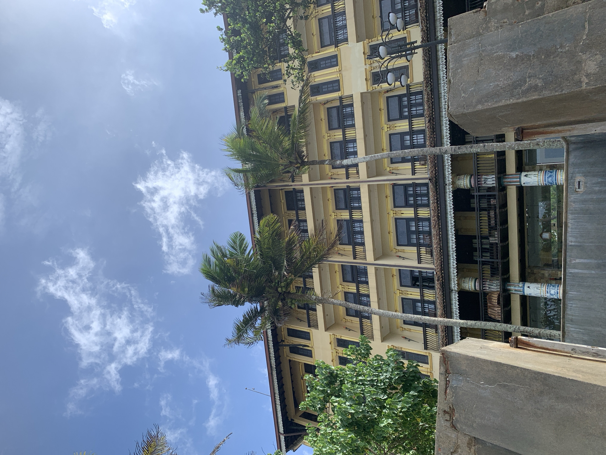 A low-angle, eye-level shot shows a large, ornate, yellow building with multiple balconies and dark-framed windows. Two palm trees with green fronds are prominently featured in the foreground, obscuring parts of the building. The sky above is blue with scattered white clouds. In front of the building, there are stone structures and a lamppost with multiple lights. The image is a photograph of the entrance of the Kathaluwa Buddhist Temple in Kathaluwa, Sri Lanka.