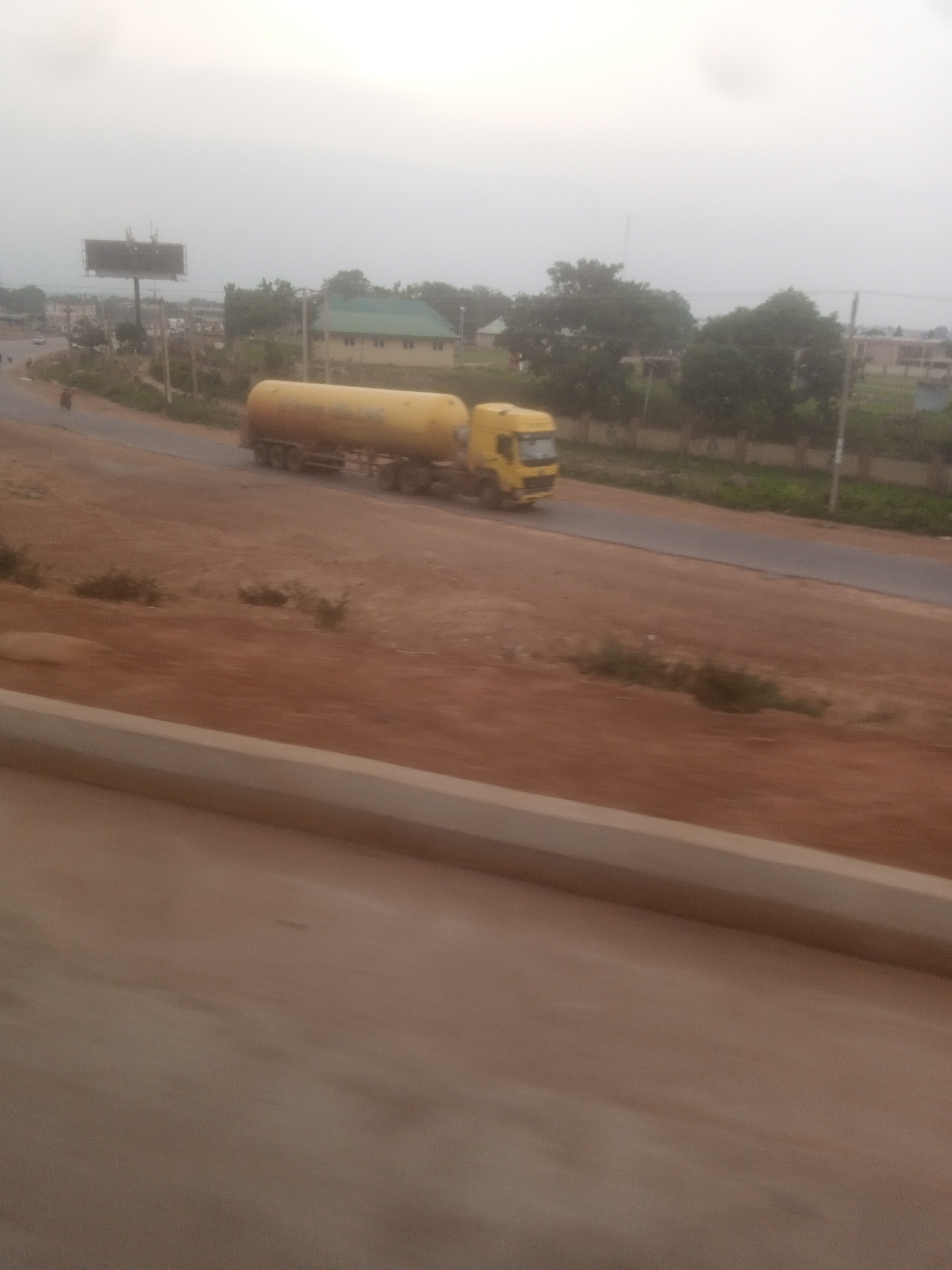 An over-exposed, hazy, daytime, eye-level, full shot depicts a yellow tanker truck driving on a paved road. The truck is positioned on the right side of the frame, angled away from the viewer and slightly to the left. The tanker is cylindrical and light yellow. The front of the truck, the cabin, is dark yellow with a black grill and white headlights. The truck has multiple wheels. The road is dark gray and appears to be a highway with no lane markings visible. The road is bordered on the left by a reddish-brown dirt embankment with some sparse vegetation. On the right side of the road, there are more trees and some buildings, including a house with a green roof and a billboard. The sky is overcast and a light gray color, suggesting either dawn, dusk, or a cloudy day. The image appears to be taken from a moving vehicle, as indicated by the motion blur in the foreground, which shows a light brown surface.