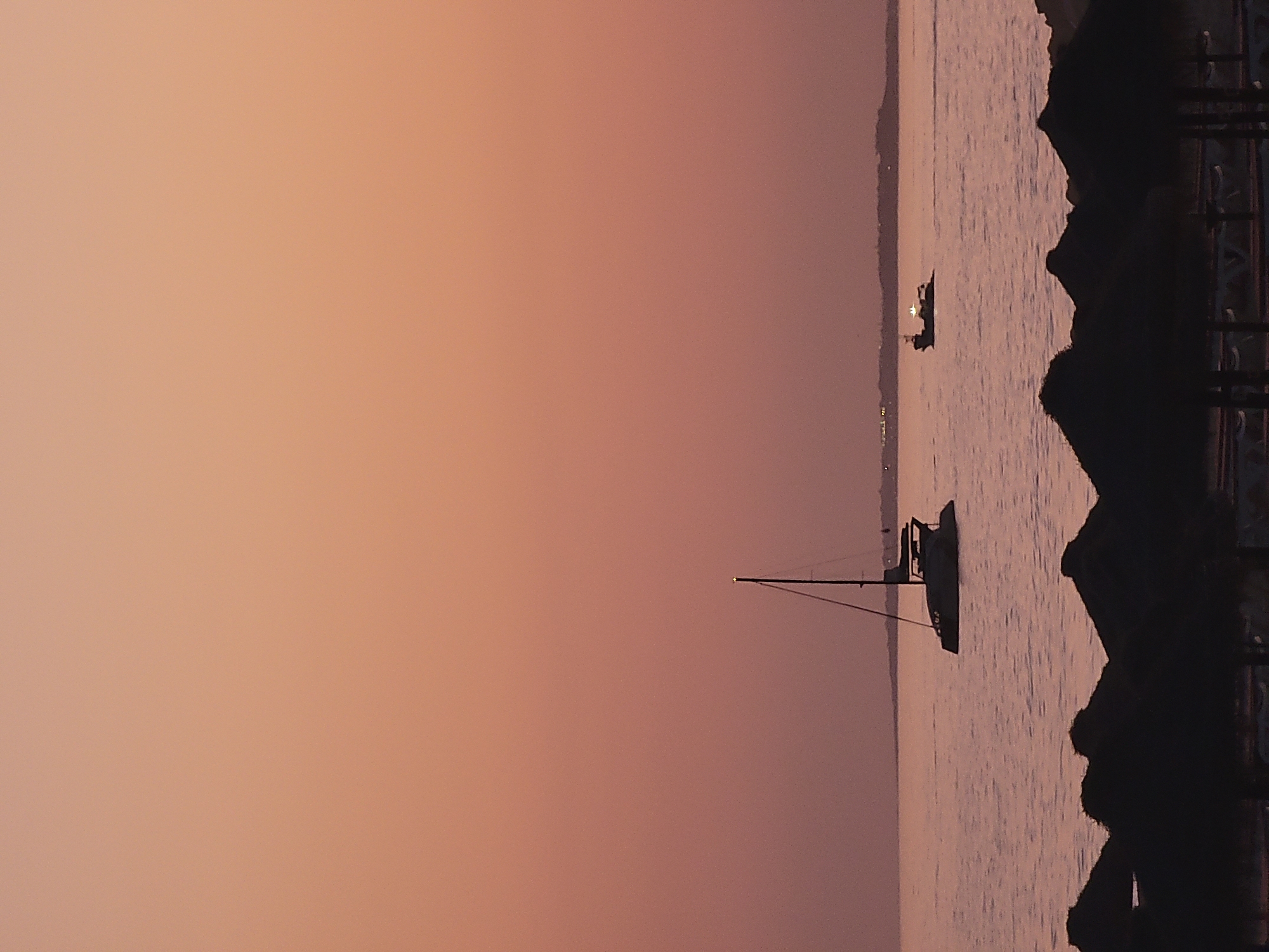 The image shows a sunset over the ocean in Quarteira, Portugal. In the foreground, several dark silhouettes of thatched umbrellas are visible. In the middle ground, a catamaran is sailing on the water, and further in the distance, a small boat with lights is visible. The sky is a gradient of soft orange and pink hues, characteristic of a sunset. The ocean is calm, reflecting the colors of the sky. The horizon line separates the sea from a faint, dark silhouette of land in the far distance.