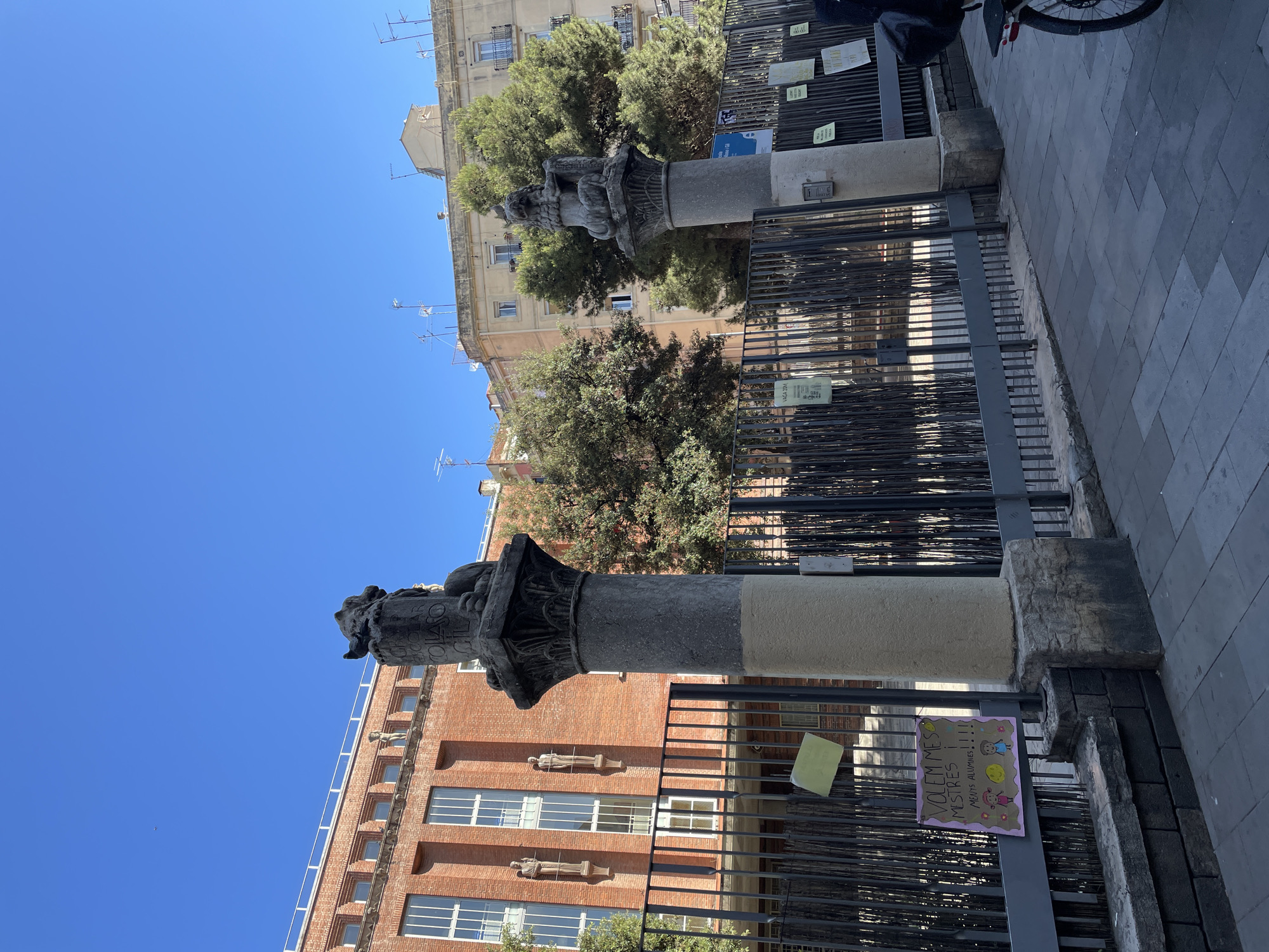 This image captures an urban street scene in Barcelona, Spain, on a bright, clear, and sunny day, evidenced by the crisp blue sky and strong shadows. The main focus is on two prominent stone columns situated behind a dark metal fence.
The column on the left features an intricately carved dark capital, possibly depicting figures or creatures, with some partially visible engraved text including "OLAO", "SOLASO", and "CHIL". The column to its right has a simpler, lighter-colored shaft. Behind these columns and the fence, green trees are visible.
In the background, two distinct buildings are seen. On the left, a multi-story brick building stands tall, characterized by rows of rectangular windows and decorative stone figures, possibly gargoyles or statues, protruding from its facade between floors. Further back, in the center, a lighter-colored building with multiple windows, balconies, and various antennas on its roof line is visible, suggesting a residential or commercial structure.
The foreground consists of a grey, patterned paved ground, likely a sidewalk or pedestrian area. A dark metal fence, composed of vertical bars, runs horizontally across the lower middle of the image, appearing to separate the paved area from the space containing the columns and trees. Attached to this fence are several paper signs. The most prominent and legible sign is handmade, pink with a purple border, and features drawings of two children and flowers. Its text reads: "VOLEM MÉS MESTRES!!!" (We want more teachers!!!) and below that, "MENYS ALUMNES !!" (Fewer students!!), indicating a protest or advocacy related to education. Other less distinct yellow and white paper signs are also affixed to the fence. A portion of a bicycle wheel and saddle bag is visible at the top right, suggesting a parked bike.