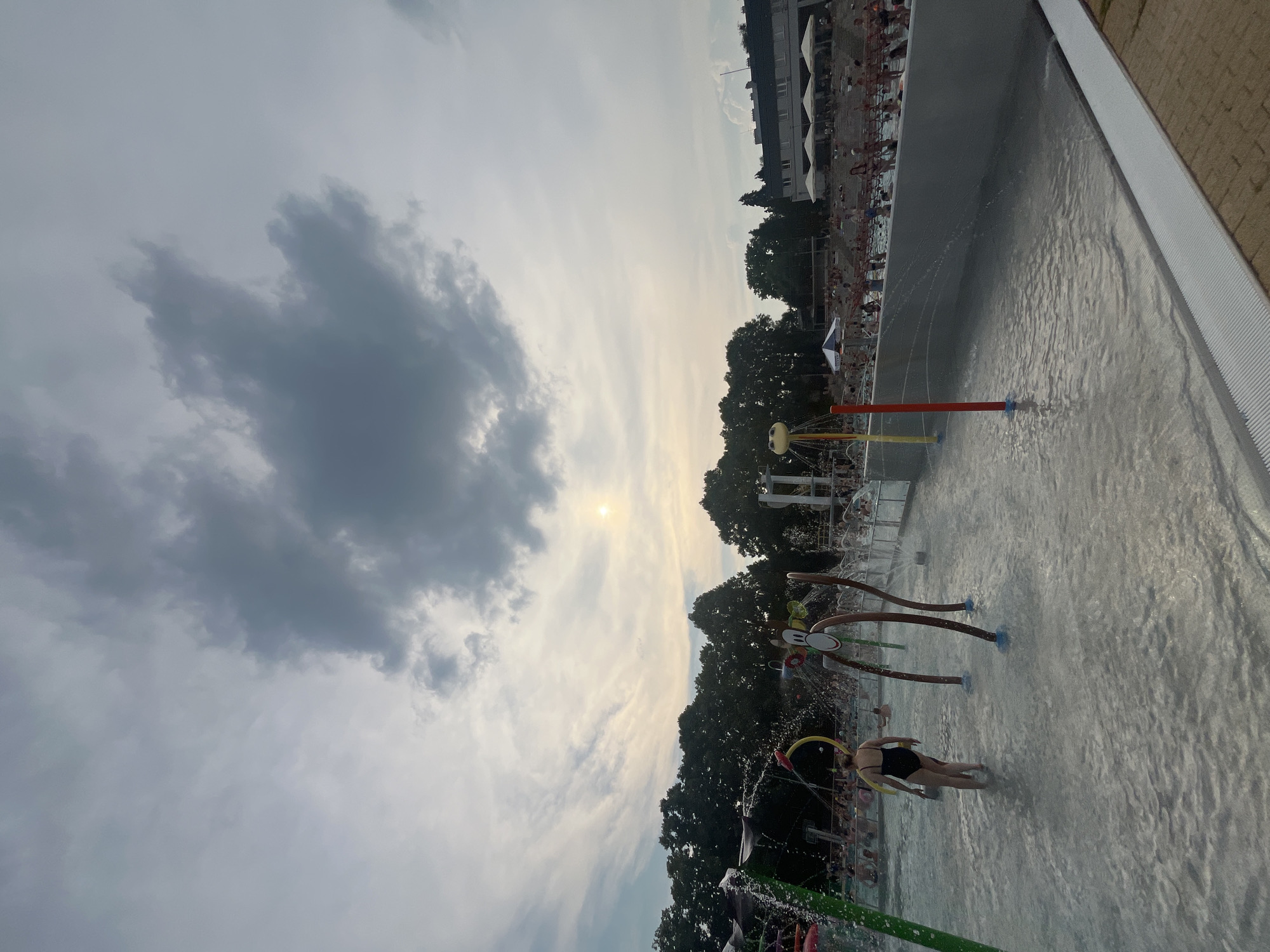 The image depicts a water park scene on an overcast day. The sun is visible through the clouds, suggesting it might be late afternoon or early evening. The main focus is a shallow pool with various water features, including sprayers and fountains, which are actively spraying water. Several people are visible in and around the pool, some wading, others interacting with the water features. One person in a black swimsuit is clearly visible in the foreground, looking away from the camera.
In the background, trees line the edge of the park, and beyond them, a building with awnings is visible, suggesting a developed area. The overall atmosphere appears relaxed and recreational. The presence of water and the clear sky, despite the cloud cover, indicates a warm weather activity. The image is captured from a low angle, looking upwards towards the sky, which is filled with dramatic clouds.