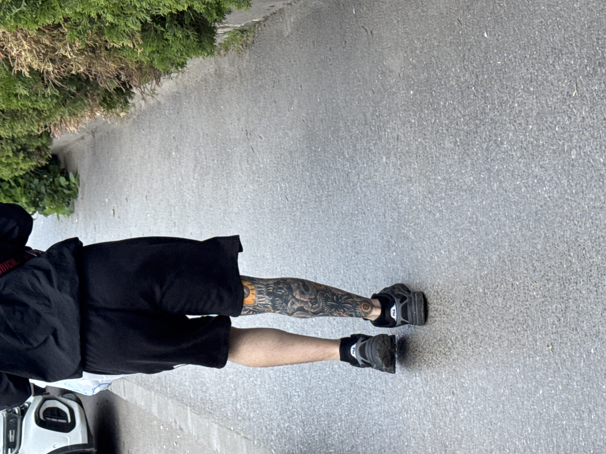 The photo shows a man from the waist down walking on an asphalt sidewalk in Sofia, Bulgaria. He is wearing black shorts, a black jacket, and black sneakers. His left leg is covered in tattoos. The sidewalk is next to a green bush. A white vehicle with black features is partially visible in the bottom left corner.