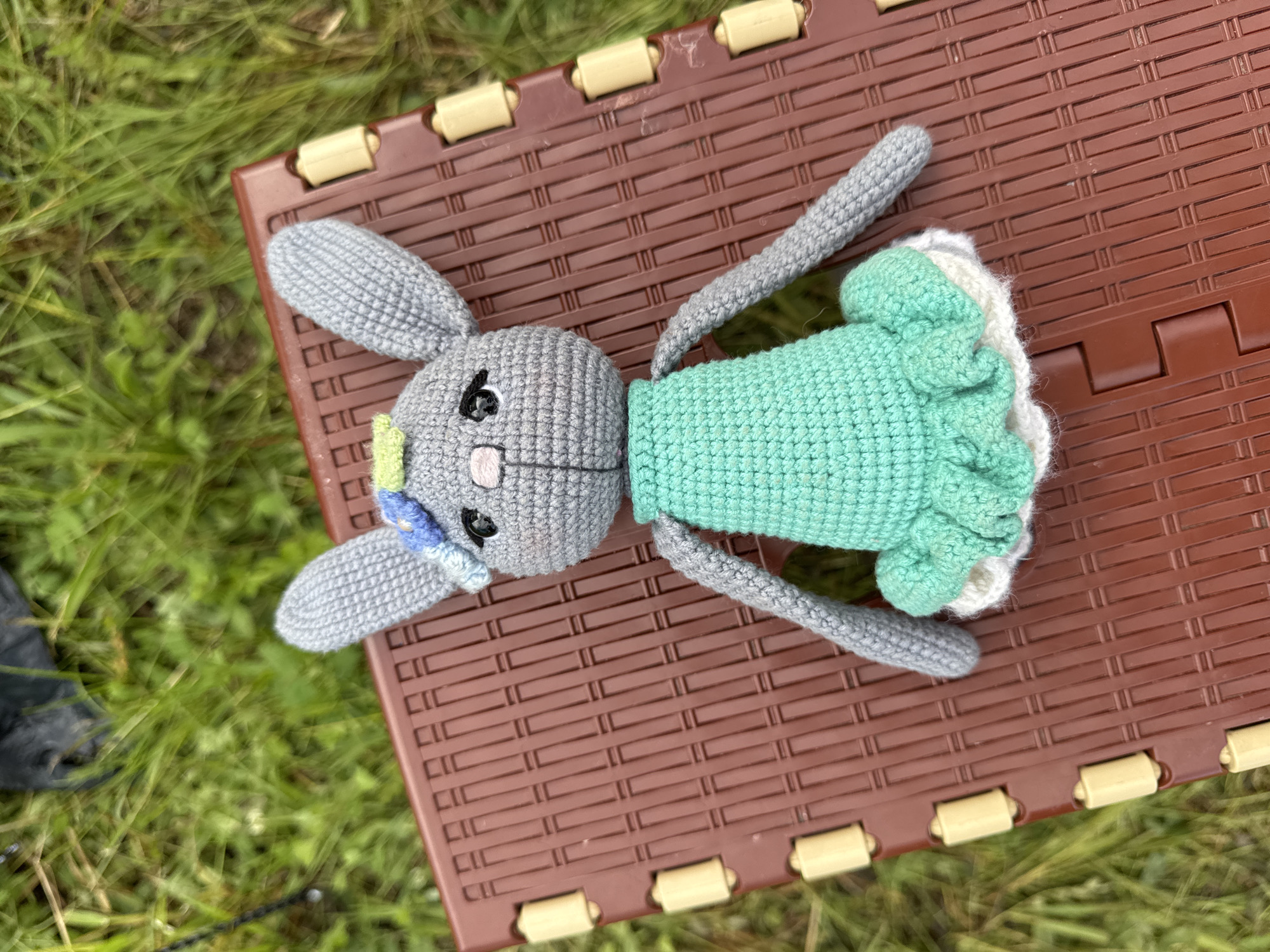 A grey crocheted bunny rabbit toy rests centrally on a brown plastic outdoor surface, possibly a folding table, with a grassy background. The bunny is light grey, features prominent ears, black embroidered eyes with white highlights, and a pink embroidered nose and mouth. It is dressed in a mint green crocheted dress with a ruffled skirt, under which a white ruffled layer is visible. A small blue and green crocheted embellishment, resembling a flower, is attached to its head between the ears. The brown plastic surface has a textured, grid-like pattern and exhibits light yellow cylindrical elements along its left edge, consistent with hinges or connectors. The surrounding environment consists of green grass, with an out-of-focus dark object and a black strap partially visible in the upper left background. The scene is located in the city of Большая Кивара, Russia.