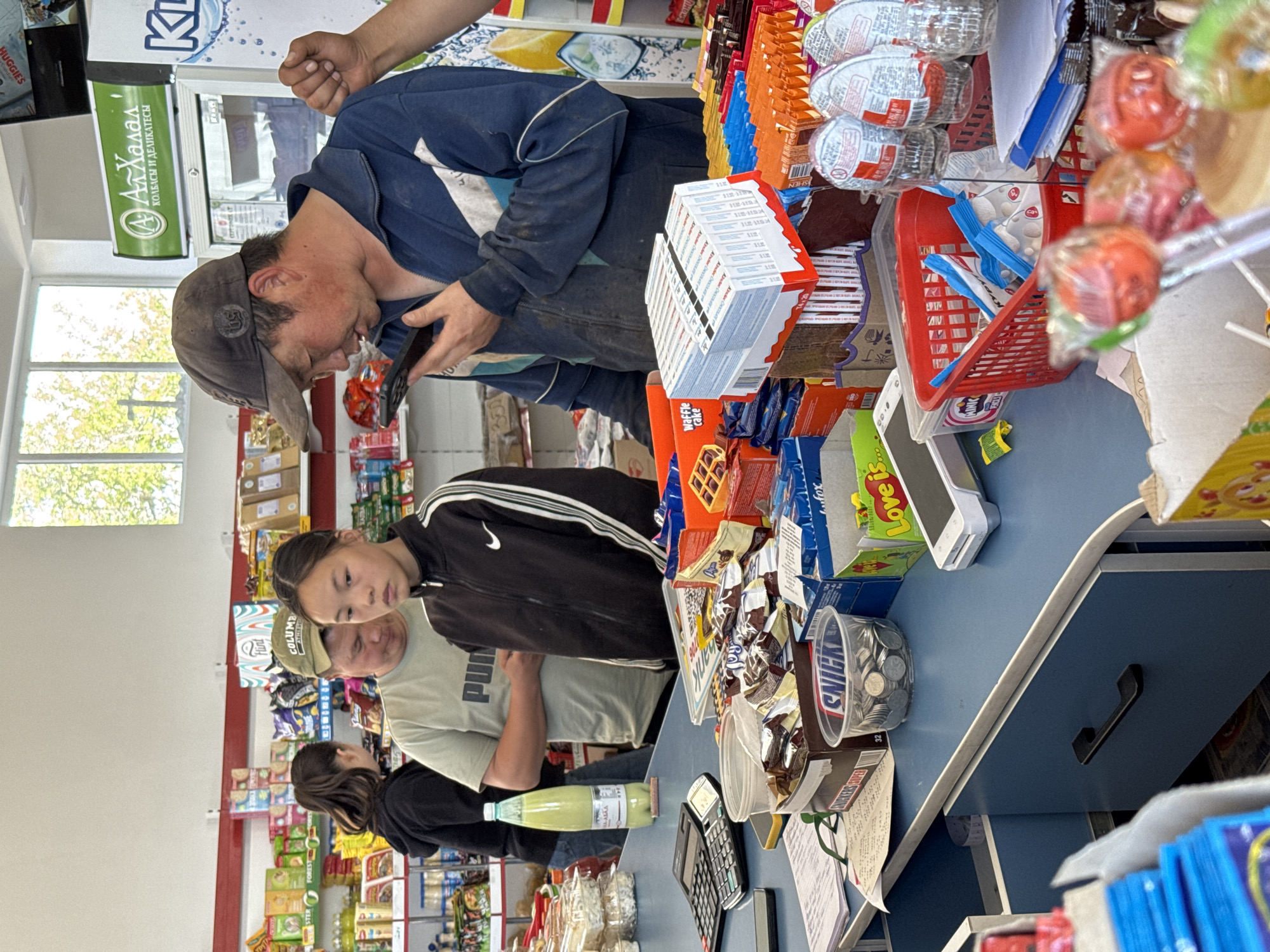 The image captures a busy scene inside a small grocery store or market in Kyzyl-Suu, Kyrgyzstan, with the camera oriented vertically. The setting features a blue counter serving as a point of transaction or display.
In the foreground, a man in a dark blue jacket and a baseball cap is intently looking at a black smartphone he holds. His hand is also visible higher up, possibly another person's arm, suggesting interaction or work at the counter. Behind him, a young woman in a black track jacket with white stripes looks towards the left, appearing to be observing or waiting. Further back, another man in a light-colored t-shirt with a "Puma" logo and a cap that says "COLUMBIA ATHLETIC" on the side is visible, along with a woman in a black top who is partially obscured.
The counter is laden with various products and store items. On the right, there are numerous stacks of "Kinder" branded items, boxes of "Waffle Cake," and other packaged snacks. A clear plastic container filled with coins labeled "SNICKERS" is also present, alongside a calculator and scattered papers, including receipts. Lollipops on sticks are displayed prominently, partially obscuring other items. Further down the counter, a light green bottled beverage is visible.
In the background, shelves are stocked with various goods, including a "HUGGIES" branded product. A refrigerator with "АлХалал КОЛБАСЫ И ДЕЛИКАТЕСЫ" (AlHalal Sausages and Delicatessen) text is noticeable, indicating specific product offerings. A window allows natural light into the store, showing trees outside, suggesting it is daytime. Other visible text includes "Love is..." and "KL" on various product displays. The overall scene depicts a typical commercial exchange or moment of activity within a local shop.