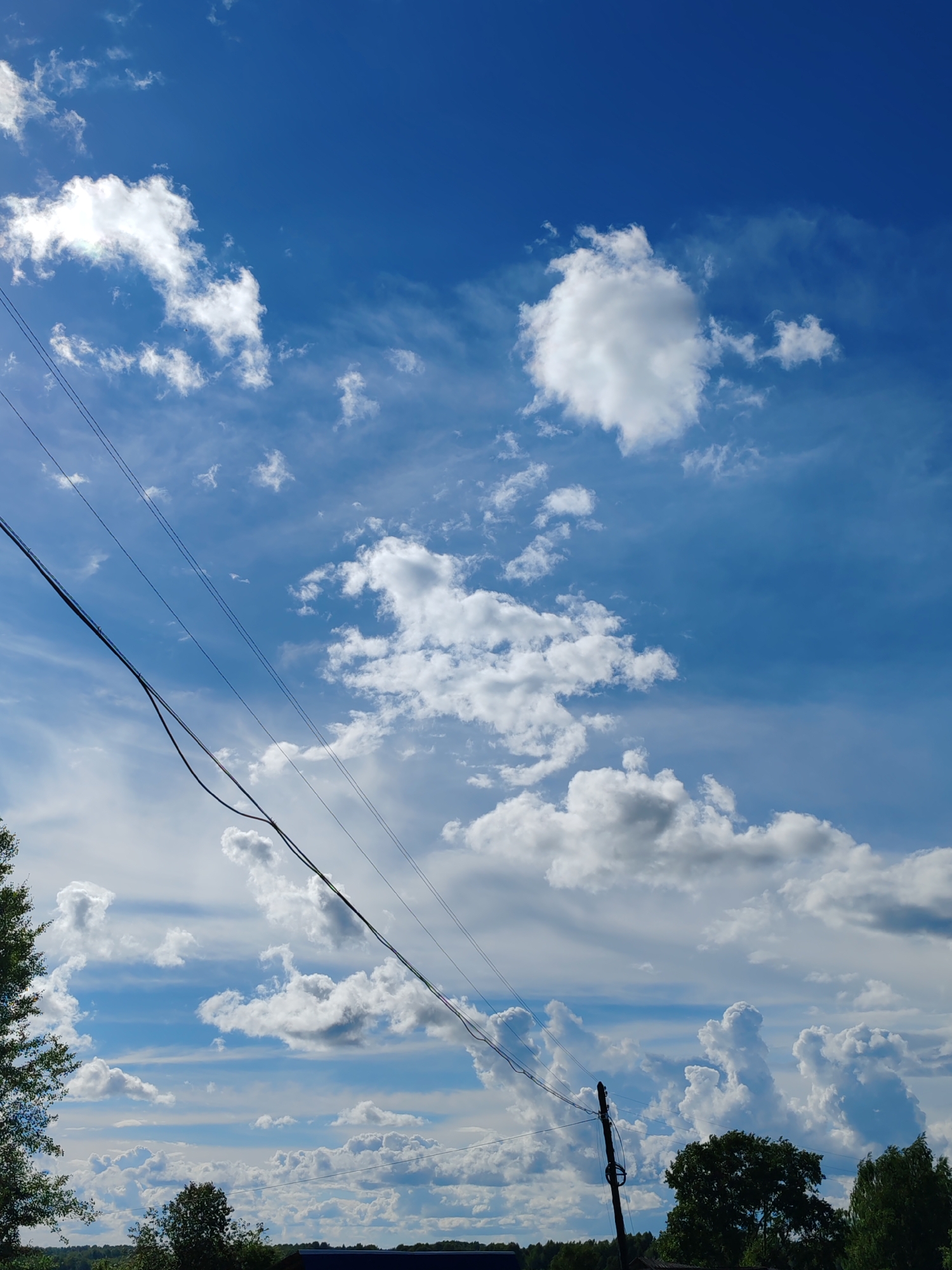 The image captures a vast expanse of a bright blue sky filled with fluffy white clouds. Multiple power lines stretch diagonally across the frame, creating a geometric contrast with the organic shapes of the clouds. In the lower portion of the image, a silhouette of trees and what appears to be the roof of a building can be seen, suggesting an outdoor, rural setting. The lighting indicates it is daytime, likely late morning or early afternoon, with the sun contributing to the bright illumination of the clouds. The scene evokes a sense of openness and tranquility, typical of a clear day in the countryside. There is no visible text in the image.