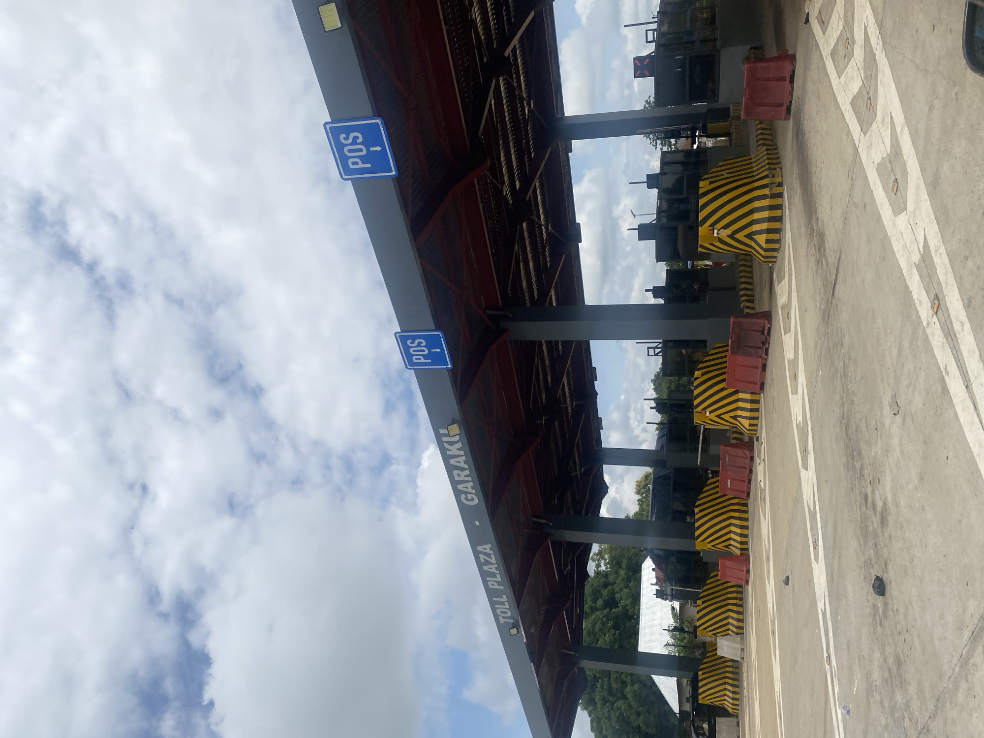A toll plaza is visible, with the words "TOLL PLAZA - GARAKU" emblazoned on the structure. Several "POS" signs with downward-pointing arrows are mounted on the overhang. The toll booths are equipped with barriers and signage. The scene is outdoors, with a cloudy sky overhead and trees visible in the background. The ground is paved with white lane markings and reflective markers. The overall environment appears to be a transportation infrastructure setting.