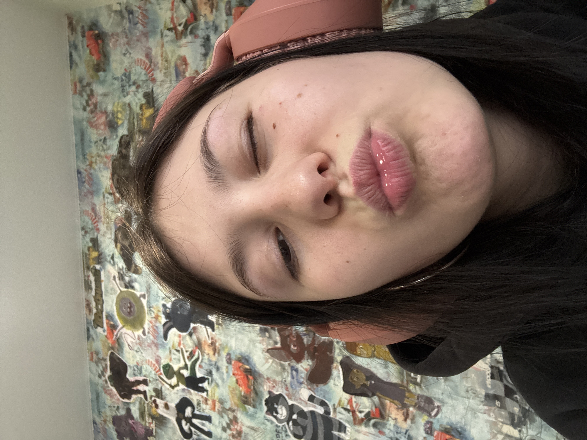 A person with dark hair and light skin is facing the camera, tilted to the left. The person has one eye closed in a wink, with puckered lips and a visible reflection suggesting lip gloss. They are wearing a pink headband. The background is a wall covered in stickers and drawings. There is no verifiable evidence to suggest the location is in the city of Фоки, Russia.