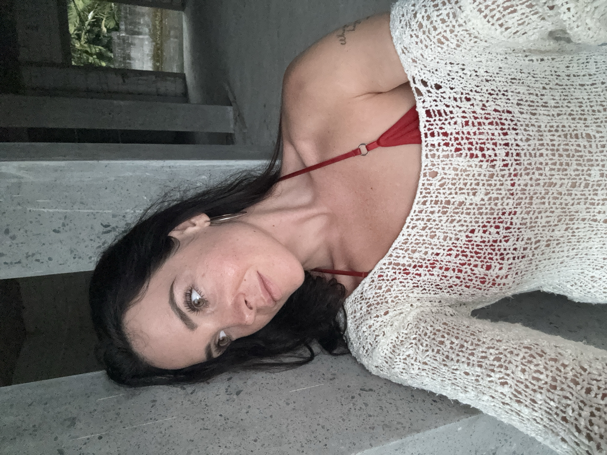 A woman with dark hair and brown eyes is leaning against a concrete pillar. She is wearing a red bikini top and a white crocheted sweater. She is looking to her left, away from the camera. The background appears to be an unfinished building with more concrete pillars and lush green foliage visible in the distance. The scene is dimly lit, suggesting it may be late in the day or indoors. The environment appears to be outdoors, possibly in a semi-developed or abandoned area, within the city of Bebandem, Indonesia.