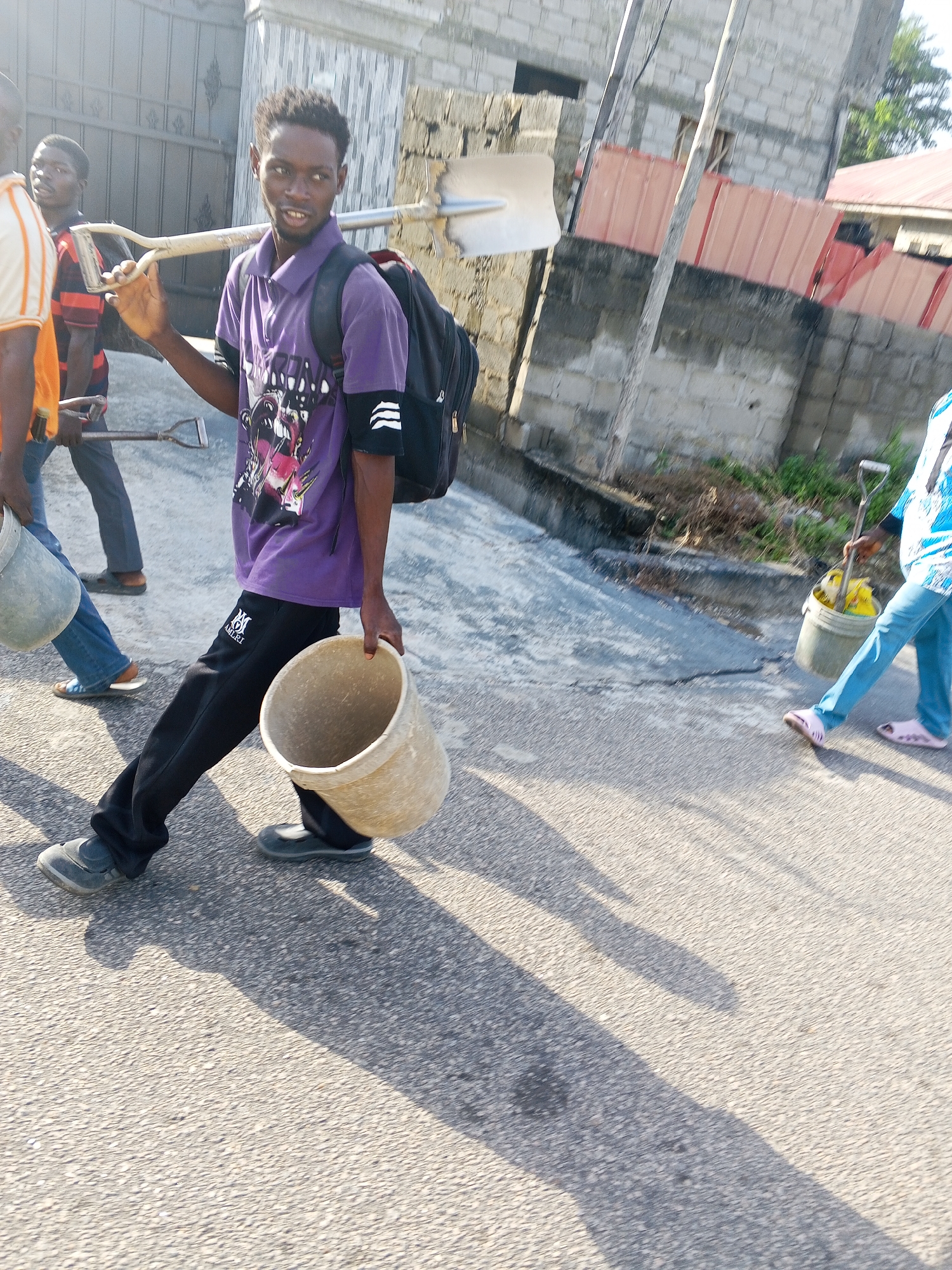 The image shows a group of individuals, likely engaged in community service or manual labor in Lokoja, Nigeria. One man in a purple shirt and black pants carries an empty bucket in his hand and a shovel over his shoulder. He wears a backpack. Other individuals are nearby, also carrying tools and buckets. The location appears to be a street in a residential or semi-urban area. The buildings in the background are constructed of concrete blocks, with some having corrugated metal roofing. The street is paved and the light suggests it is daytime.