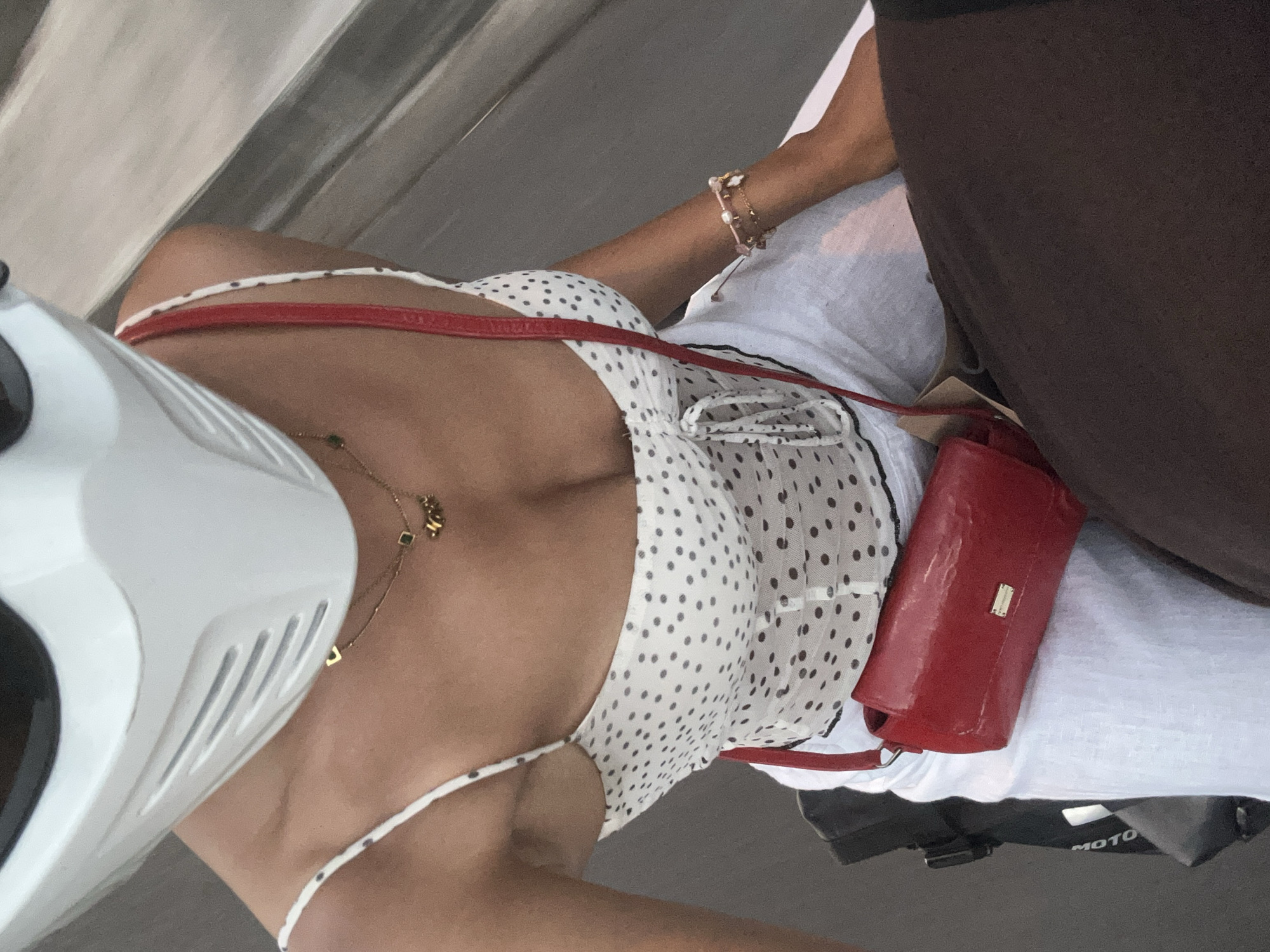 A woman wearing a white full-face motorcycle helmet, a white and black polka dot camisole, white pants, and a red crossbody bag rides on the back of a motorcycle in Ubud District, Indonesia. Her left arm, adorned with a bracelet, is visible resting on her lap. The scene appears to be an outdoor setting, likely a street in Ubud, with a blurred background suggesting movement.
