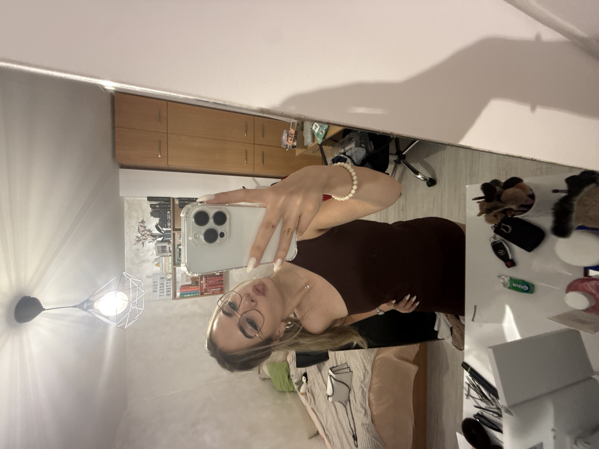 A woman is taking a selfie in a mirror. The reflection shows a room with a bed, dresser, desk, and various items scattered on the desk. The location is not specified within the image and cannot be determined as Blagoevgrad, Bulgaria.