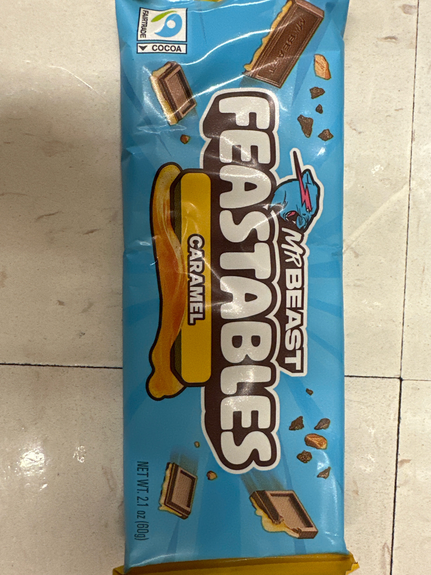 A bright blue wrapper with the text "FEASTABLES" and "CARAMEL" is laid flat on a cream-colored tiled floor. The wrapper features images of chocolate pieces and caramel. A portion of a yellow wrapper is visible at the bottom of the image. The lighting suggests an indoor environment, possibly a kitchen or pantry, with shadows indicating overhead light sources. The context of Camp Hill, United States of America is not discernible from the image itself.