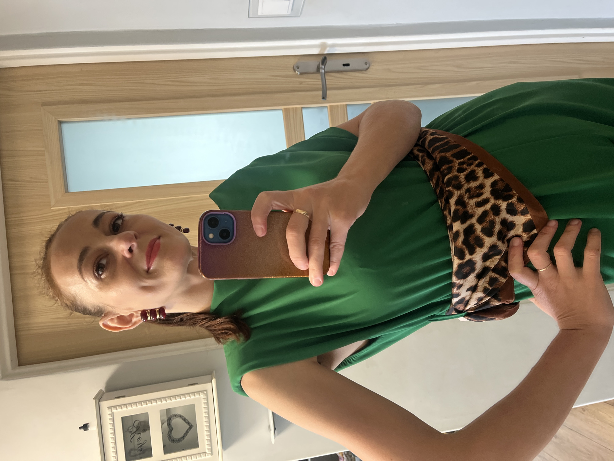 A woman is taking a selfie in a mirror. She is wearing a green dress with a leopard print scarf tied around her waist. She has on large earrings and a ring on her left hand. The background shows a door and a framed picture on the wall.