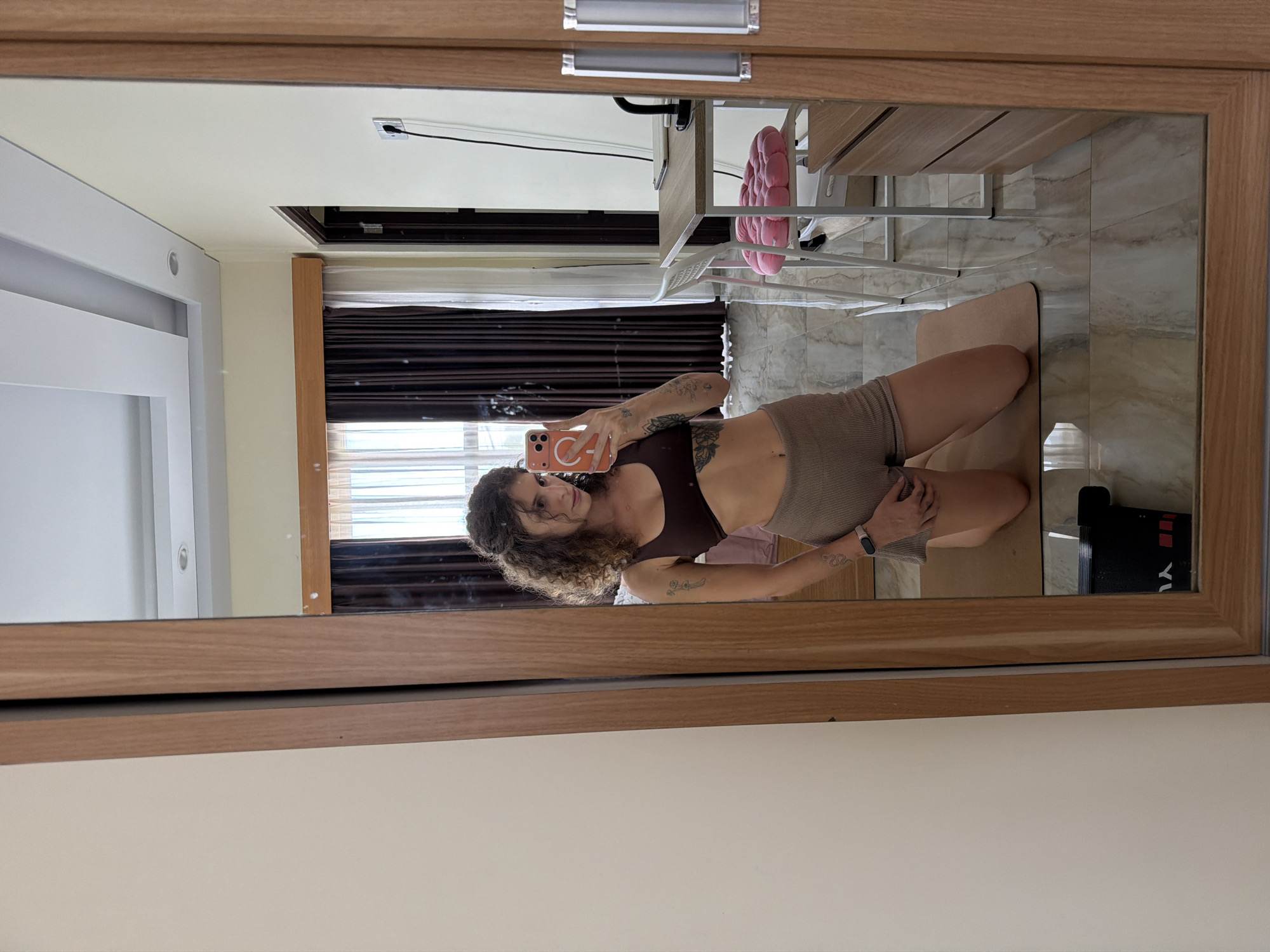 A young woman with curly brown hair is captured in a mirror selfie, kneeling on a beige yoga mat. She is wearing a dark brown sports bra and light brown ribbed shorts, with visible tattoos on both arms and a smartwatch on her left wrist. She holds a peach/orange smartphone, featuring a MagSafe ring, up to take the photo, and has a subtle, relaxed smile.
The setting appears to be a bright indoor room in North Kuta, Indonesia, likely a personal space like a bedroom or living area. The room has light-colored walls, a tiled floor with a marble-like pattern, and light wood-toned furniture, including a desk and a chair with a pink fluffy cushion. A window with blinds is visible in the background, letting in ample natural light, indicating it is daytime. Other details include a wall outlet, a white door, and a black object on the floor near the yoga mat with partially visible white text, possibly "YA". The overall atmosphere is relaxed and comfortable.