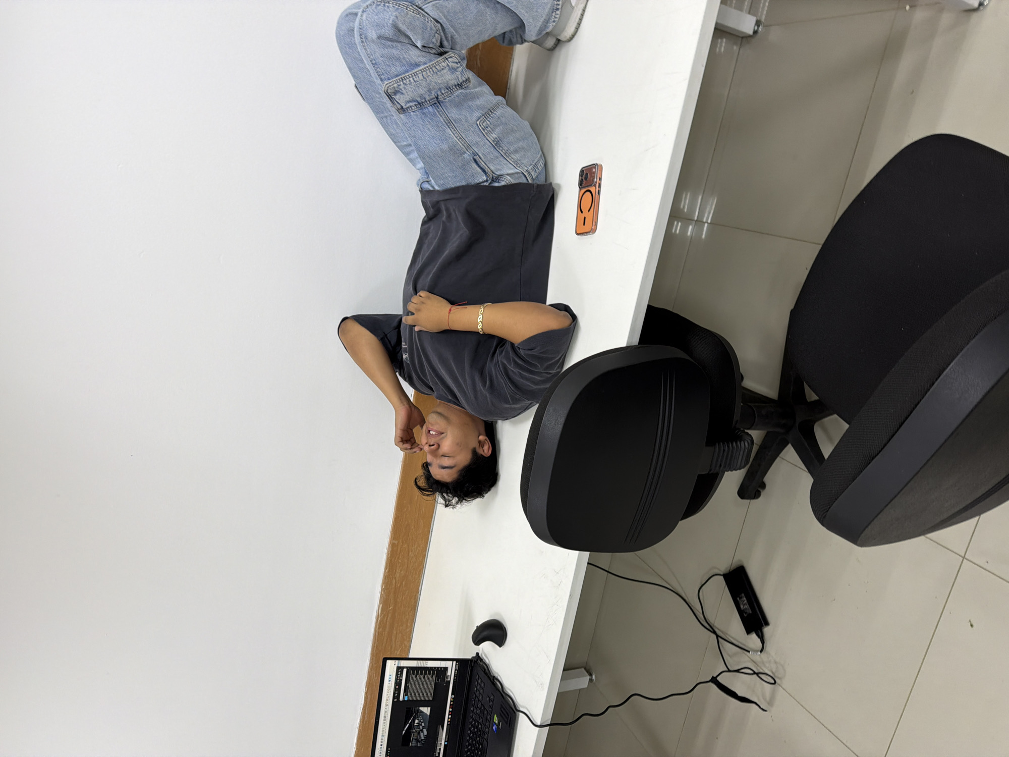 A person is lying on a white desk in San Andrés Cholula, Mexico. They are wearing a grey t-shirt and light blue jeans. A black laptop, black mouse, and cell phone are on the desk. A black office chair sits next to the desk. A charging cord is on the tiled floor. The walls are white.