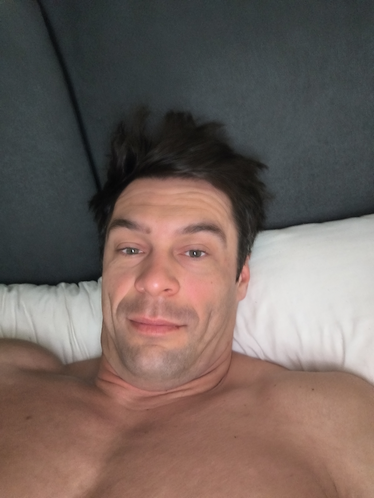 A shirtless man is lying on a bed with a white pillow and a grey headboard. He is looking directly at the camera. The scene's location cannot be determined from the image.