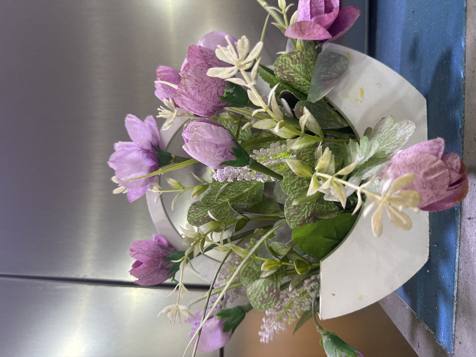 A floral arrangement in a white, U-shaped vase sits on a blue surface. The vase contains artificial flowers and greenery, predominantly in shades of pink and purple, with some lighter accents. The background is a metallic, brushed surface. The scene appears to be indoors, possibly a domestic or commercial setting in Shofirkon, Uzbekistan.