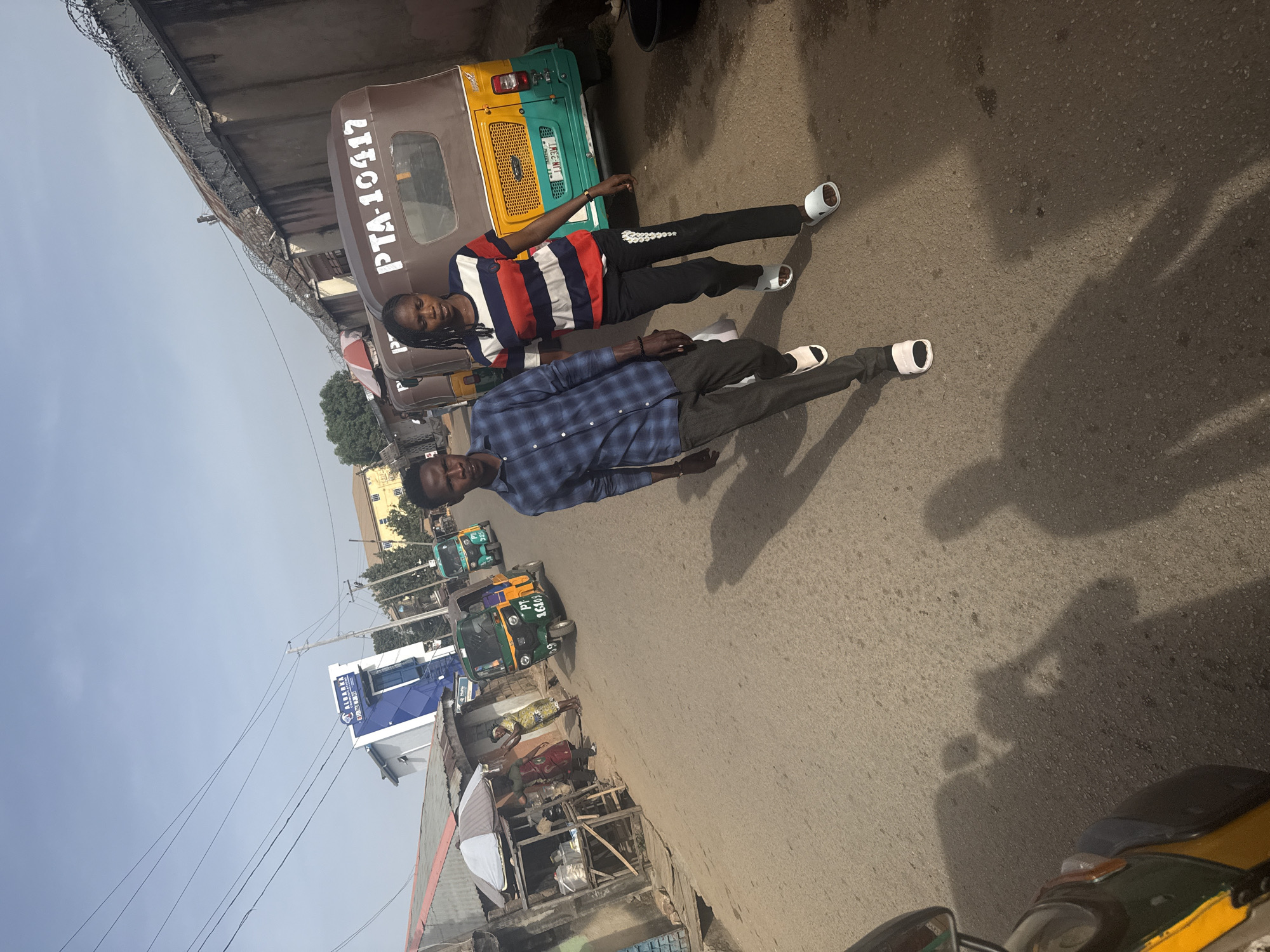 Two people are lying on a street in Jos, Nigeria. One person wears a blue and black checkered shirt, gray pants, and white slide sandals. The other wears a striped red, white, and blue shirt, black pants, and white slide sandals. Several three-wheeled vehicles called "keke napep" are parked along the street, and a building with a blue and white sign can be seen. The sky is overcast. A barbed wire fence overhangs a bridge above the road. The number "PTA-10417" is printed on the side of one of the vehicles.
