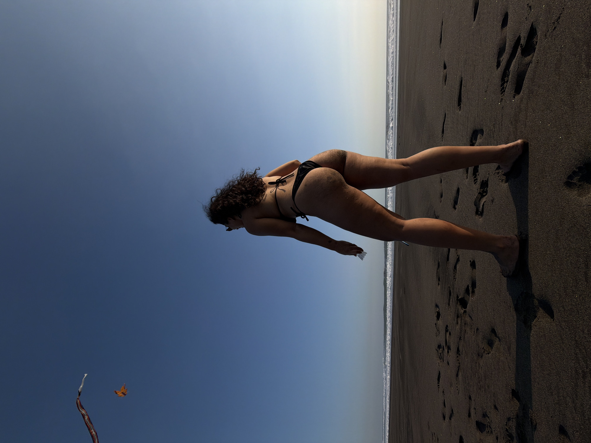 On a dark sand beach in Selemadeg, Indonesia, a person is observed from behind, facing the ocean under a clear blue sky. The individual is a female with dark, curly hair, wearing a black string bikini and sunglasses. She stands barefoot on the black, volcanic sand, which shows numerous footprints. Sand is visible on her left buttock and thigh. Her left hand holds a small white object. Gentle ocean waves break in the distance along the horizon. In the upper left portion of the sky, a colorful streamer and an orange, butterfly-shaped kite are airborne. The lighting suggests either late afternoon or early morning, with the sky appearing brighter towards the horizon on the right.