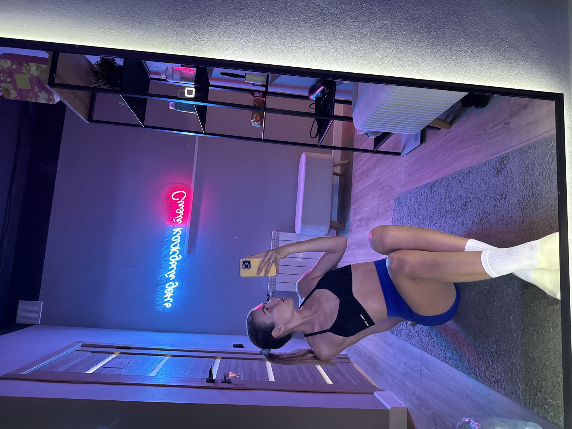 A young woman is captured in a mirror reflection, lying on a gray rug. She's dressed in a black sports bra and blue shorts, with white socks. In her right hand, she holds a yellow smartphone as if taking a selfie. The background consists of a gray wall, a wooden door on the left, and a shelving unit filled with various items on the top, along with a bench. A neon sign is visible on the wall. The location appears to be inside a building.