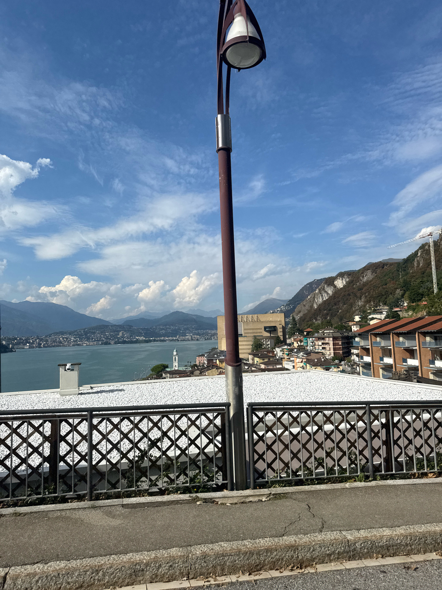 The image depicts a view of Campione d'Italia, Italy, with Lake Lugano and mountains in the background. A street lamp stands prominently in the foreground, partially obscuring the sky. The street lamp is dark brown with a metal housing for the light. Below the lamp, a metal railing with a diamond pattern separates the viewpoint from the buildings and the lake. The buildings in the mid-ground include a white-roofed structure with a chimney, residential buildings with red-tiled roofs, and a tall, modern beige building. A church steeple is visible among the buildings near the lake. The lake itself is a calm body of water, reflecting the blue sky and white clouds. In the far distance, rolling hills and mountains are visible, some with rocky faces and others covered in trees. A construction crane is visible on the hillside to the right. The sky is a bright blue with scattered white clouds, some of which are large and puffy. The ground in the immediate foreground is asphalt with a stone curb. The overall scene is one of a sunny day in a scenic, lakeside Italian town.