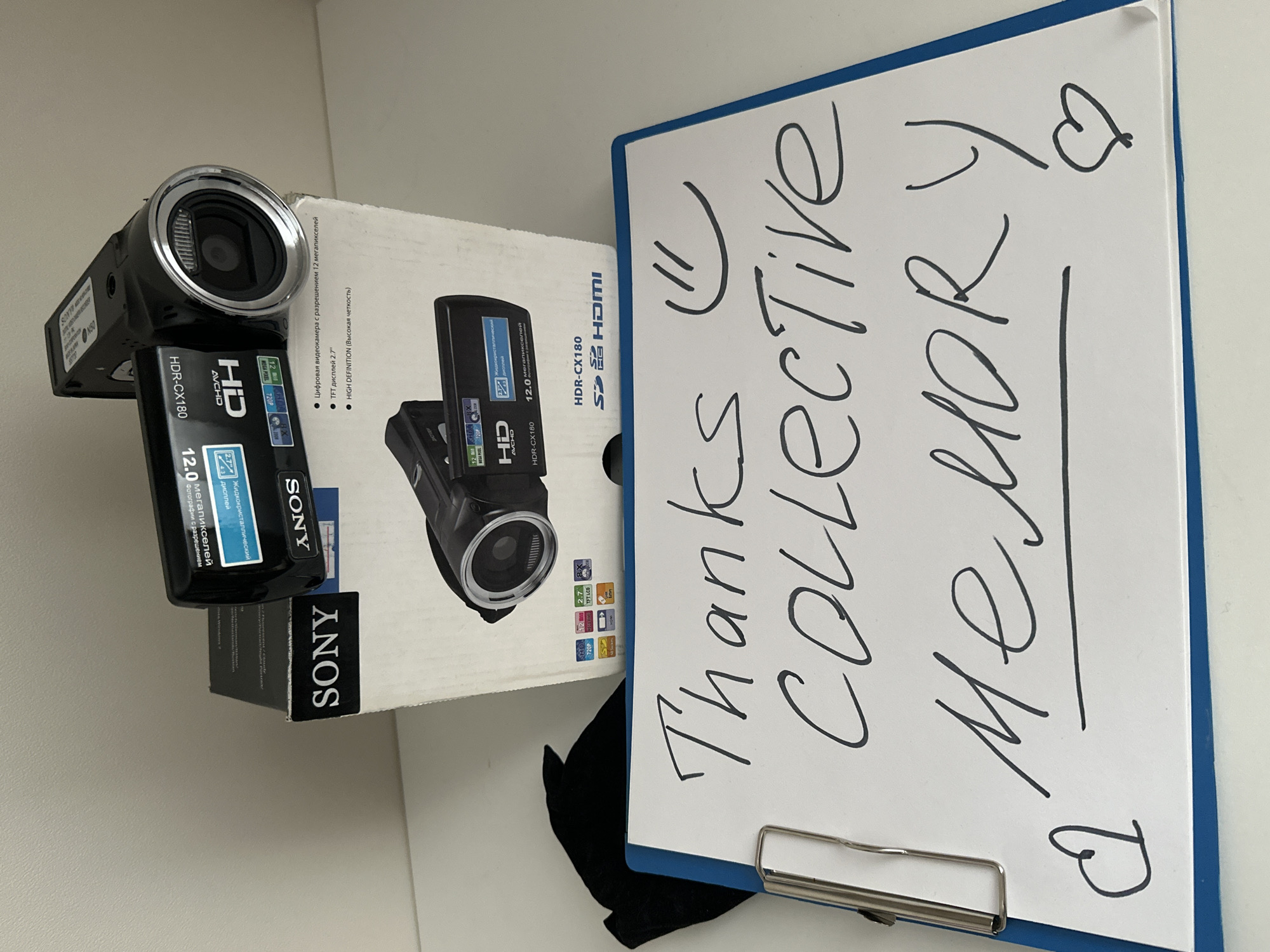 An overhead shot captures a black Sony HDR-CX180 camcorder resting on its white retail box, next to a white clipboard with a blue frame. The setup is arranged on a light-colored surface, possibly a desk, against a plain, light-colored wall.
The camcorder itself is prominently displayed, showing its lens and several labels including "HDR-CX180," "HD," "AVCHD," and indicating "12.0 мегапикселей" (12.0 megapixels) and "720P." The accompanying box is white with an image of the camcorder and various technical specifications. Visible text on the box, partly in Russian, confirms "HDR-CX180," "HD," "AVCHD," "12 MEGAPIXEL," "2.7 TFT LCD," "SD," and "HDMI" compatibility. Russian text reads: "Цифровая видеокамера с разрешением 12 мегапикселей" (Digital camcorder with 12 megapixel resolution), "TFT дисплей 2.7"" (2.7" TFT display), and "HIGH DEFINITION (Высокая четкость)" (High Definition (High Clarity)).
The white clipboard contains a handwritten message in black marker: "Thanks Collective MEMORY," accompanied by a smiley face and a heart icon. The word "MEMORY" is underlined. A black fabric item is visible underneath the clipboard, possibly a cloth or a small bag. The overall impression is a display of a new camcorder, possibly a gift or purchase, with a heartfelt thank you note, suggesting its purpose for capturing shared moments. The Russian text on the box supports the Moscow, Russia location context.