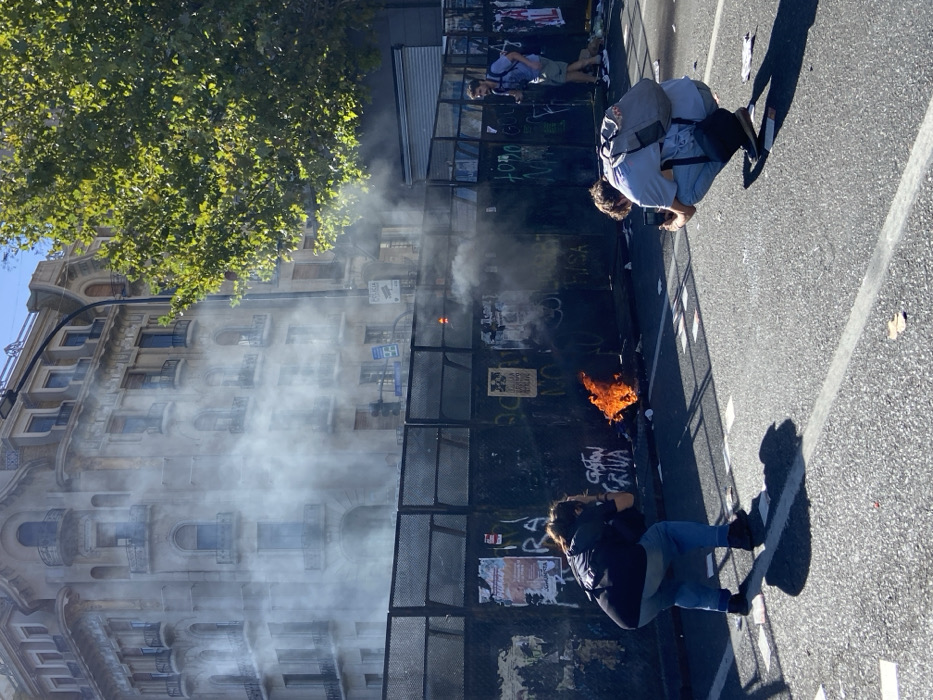 The image captures an urban scene in Buenos Aires, Argentina, dominated by an active fire and smoke. In the immediate foreground, an asphalt street with white markings is littered with papers and debris. Two individuals are crouched on the street, facing away from the viewer, actively photographing a burning object with mobile phones. The person on the left wears a dark top and blue jeans, while the person on the right wears a light-colored shirt, blue jeans, and a grey backpack. Both cast long shadows onto the street, indicating strong sunlight.
Mid-ground features a dark, temporary metal fence or barricade, adorned with various posters and graffiti, including "GASTON GRIMA." Behind the left side of this fence, a localized fire burns on the ground, emitting significant white and grey smoke that billows upwards, partially obscuring the background. A "POLICIA" sign is visible on a pole behind the fence, towards the right.
In the background, a multi-story building with classical architectural elements, including ornate balconies and arched windows, is visible, partially veiled by the rising smoke. To the far right, a large tree with green foliage partially obstructs the view of the background building. Further to the right, behind the fence, a third individual wearing a light shirt, shorts, and a backpack is seen moving.