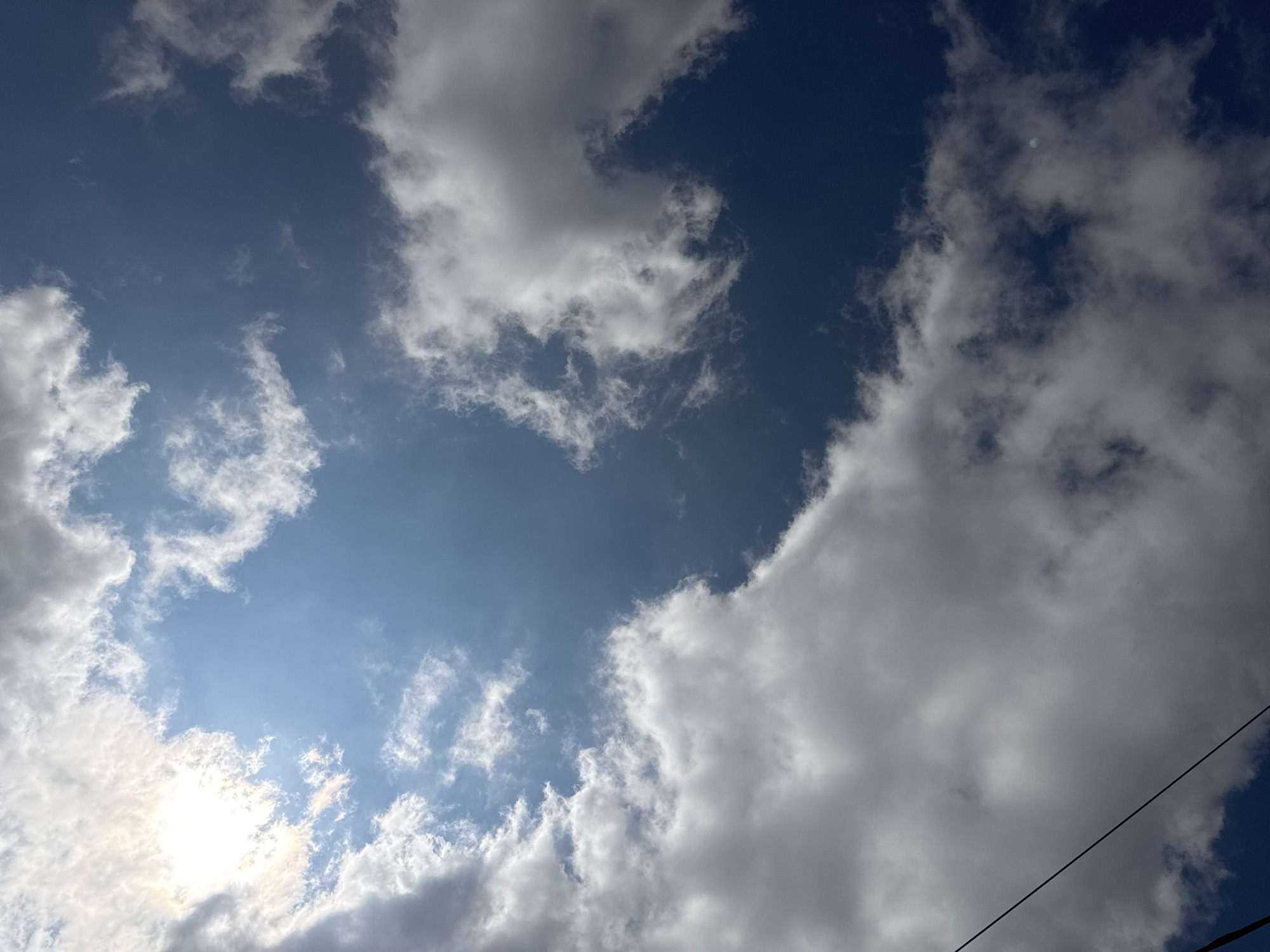 The image is an upward-facing shot of a partly cloudy sky on a bright day. The sun is visible in the upper left corner, partially obscured by white, fluffy clouds. The clouds are scattered throughout the sky, with some appearing thin and wispy and others more dense and voluminous. The sky is a deep blue color. A thin, dark wire runs diagonally from the bottom left towards the center of the frame. The image was taken in Nikolaevka, Russia.