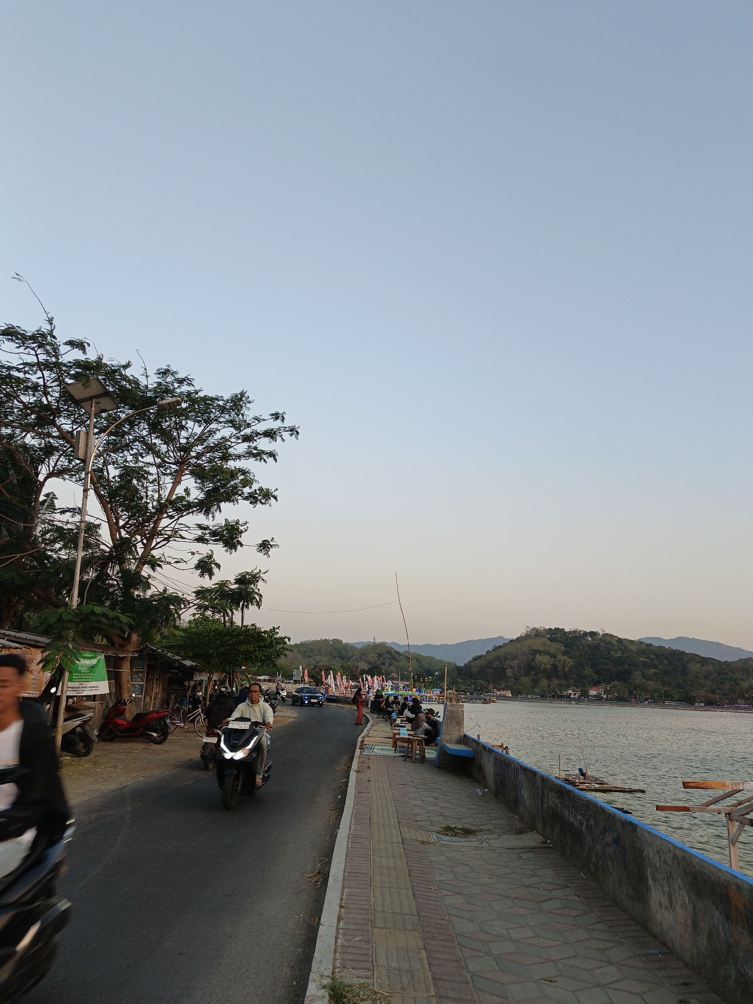 The image depicts a seaside road in Krakitan, Indonesia, at dusk. The sky is a pale, clear blue, transitioning to a soft orange near the horizon. On the left side of the road, a tall tree with sparse foliage is illuminated by a solar-powered streetlamp. Several small shops and kiosks are visible beneath the tree, with motorcycles parked in front. A man is riding a scooter on the road, and a blur of another person on a scooter is in the foreground.
The road runs parallel to a paved walkway which borders a body of water. Along the walkway, a concrete barrier with a blue trim separates the path from the water. Several people are seated at tables on the walkway, facing the water. A small raft or floating platform is visible in the water. In the distance, rolling hills covered in lush vegetation form the backdrop, with some houses nestled among them. The overall scene suggests a tranquil evening ambiance in a coastal town.