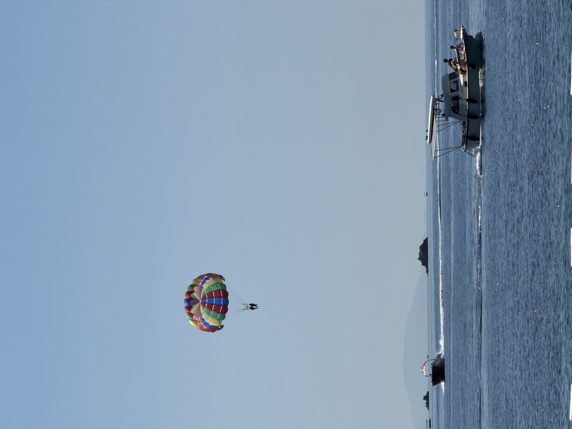 This image depicts a sunny maritime scene near Budva, Montenegro, under a clear, light blue sky. In the upper center, a brightly colored parasail, segmented with red, green, blue, yellow, and orange stripes, suspends two silhouetted individuals high above the water. The sea below is a deep blue, exhibiting gentle ripples and sunlight reflections. Two motorboats are navigating the water. On the left, a dark-hulled speedboat travels towards the left, generating a distinct white wake. On the right, a lighter-colored cabin cruiser moves rightward; it features a surfboard mounted on its roof canopy, black fenders along its side, and at least three individuals are visible on its deck. In the distant background, several dark, rocky islets are present, with a faint, hazy light blue-grey landmass extending along the horizon.