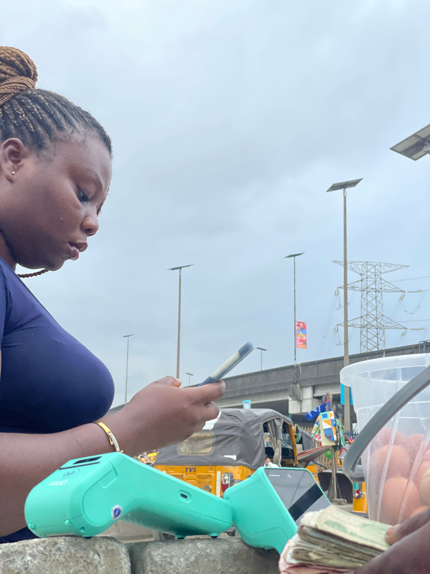 In Ajah, Nigeria, under a cloudy sky, a Black woman with braided hair tied in a bun is in the foreground, facing left and looking down at her smartphone. She wears a dark blue top and a delicate gold bracelet on her left wrist. To her right, a teal-colored credit card machine rests on a rough stone surface. In the background, several yellow auto-rickshaws, known as "keke napeps," are parked. Beyond them, a large electrical pylon and multiple streetlights with solar panels are visible, suggesting an urban or semi-urban environment. A transparent plastic bucket filled with eggs is also present on the right side of the frame. The scene captures a moment of transaction, possibly at a market or roadside stall, amidst the bustling activity of Ajah.