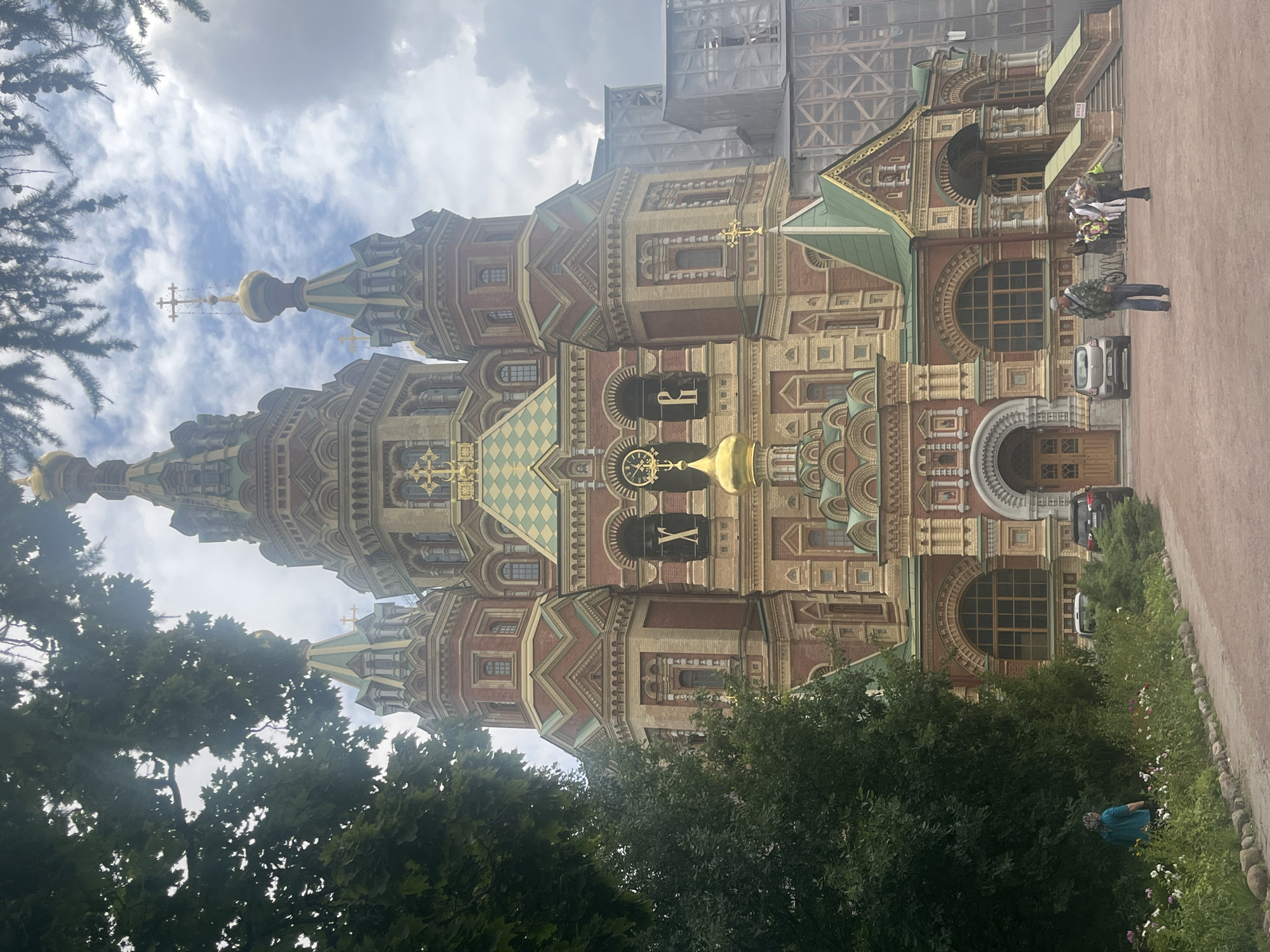 The image shows the Peterhof Grand Cathedral, an Orthodox church located in Peterhof, Russia. The cathedral is a large, ornate building with multiple golden domes and intricate brickwork. The facade is decorated with crosses, intricate carvings, and decorative arches. In front of the cathedral, there is a paved area with some trees and flowerbeds. Several people are visible in the scene, some walking on the paved area, while others are near the entrance of the cathedral. There are also a few cars parked in the vicinity. The sky is partly cloudy, and the overall lighting suggests it is daytime.