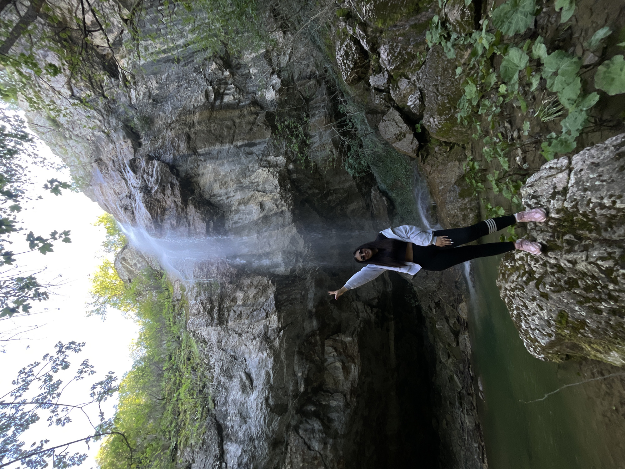 A woman stands on a rock next to a waterfall in Veselinovo, Bulgaria. She wears a grey hoodie, black leggings, and pink shoes. The waterfall cascades down a rocky cliff face covered in moss and greenery. The water collects in a dark pool at the bottom. Trees and vegetation frame the top and sides of the scene.