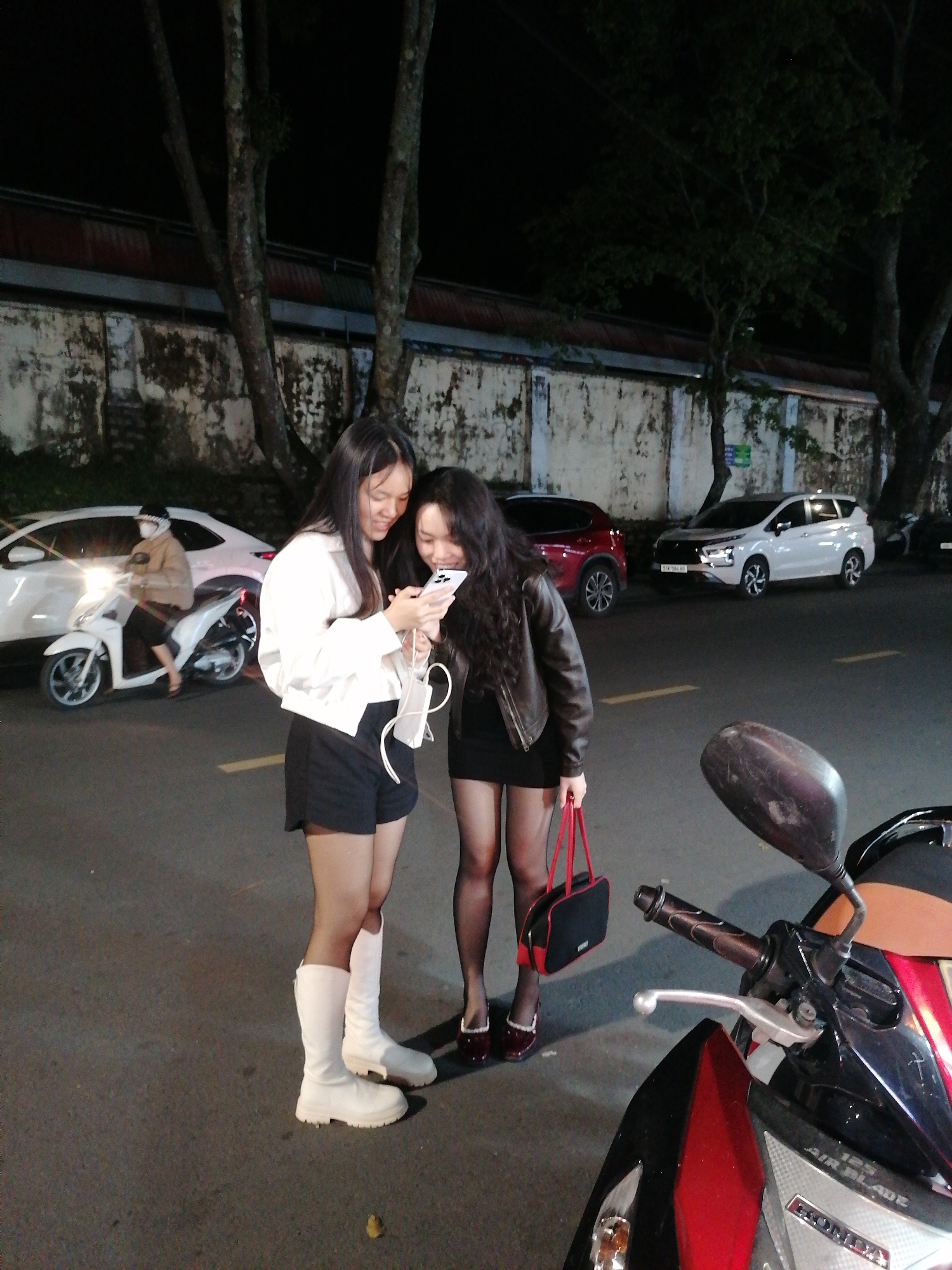 In an outdoor urban setting at night, likely in Da Lat, Vietnam, two young women are standing on an asphalt road. The woman on the left, with long dark hair, wears a white long-sleeved top, black shorts, and tall white boots. She holds a white mask with cords hanging from it. The woman on the right, also with long dark hair, wears a dark leather jacket, a short black dress, black pantyhose, and dark red heeled shoes. She carries a small red and black handbag. Both women are looking at a smartphone held by the woman on the left.
In the background, a white car is parked on the right side of the road. Further back, a wall with a weathered texture and some greenery is visible. Trees with visible bark and sparse foliage line the background. A white scooter with a rider wearing a light-colored jacket and a face covering is in motion on the left side of the road, with its headlight illuminated. The ground is a dark asphalt surface with painted yellow lane markings. The overall scene suggests a casual nighttime interaction on a street.