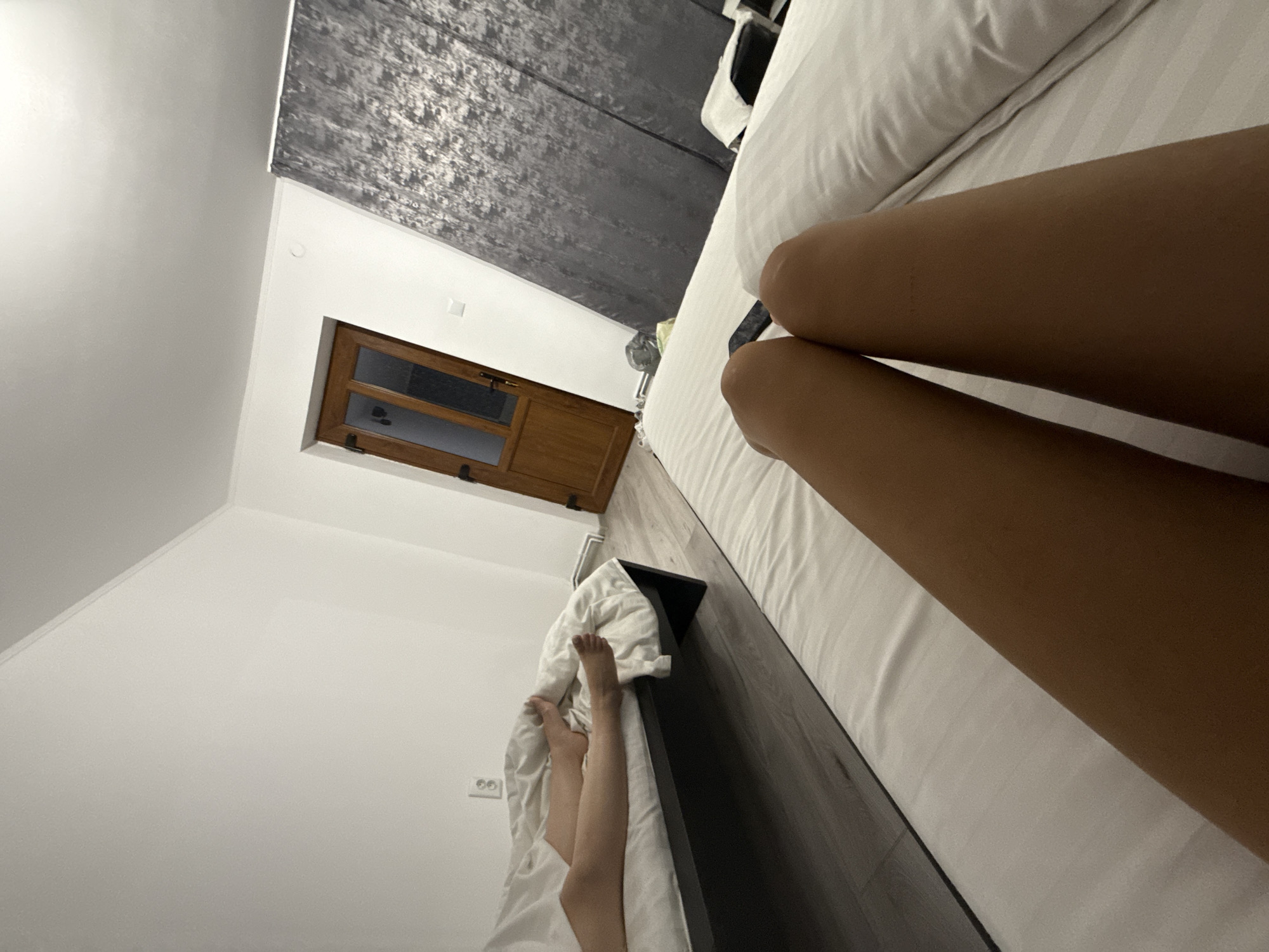 The image shows a bedroom interior, likely in Cârțișoara, Romania. In the foreground, a person's legs are visible, lying on a bed with white striped bedding. The viewpoint is from the perspective of someone lying down. In the mid-ground, another bed is partially visible with a person's legs and feet covered by white bedding. The room features light-colored walls and a wooden door with glass panels. A gray, textured curtain hangs on the wall opposite the beds. The flooring appears to be dark wood laminate. The lighting suggests an indoor setting, possibly evening or with artificial light.