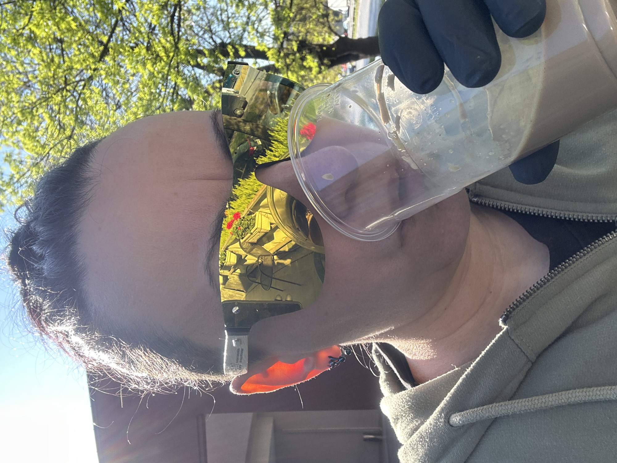 A person wearing sunglasses and a hooded sweatshirt is drinking from a clear plastic cup containing a light brown liquid. They are also wearing black gloves. The sunglasses reflect an outdoor scene of greenery and possibly some structures. The background includes trees with green leaves and a clear blue sky. The location cannot be determined from this image.