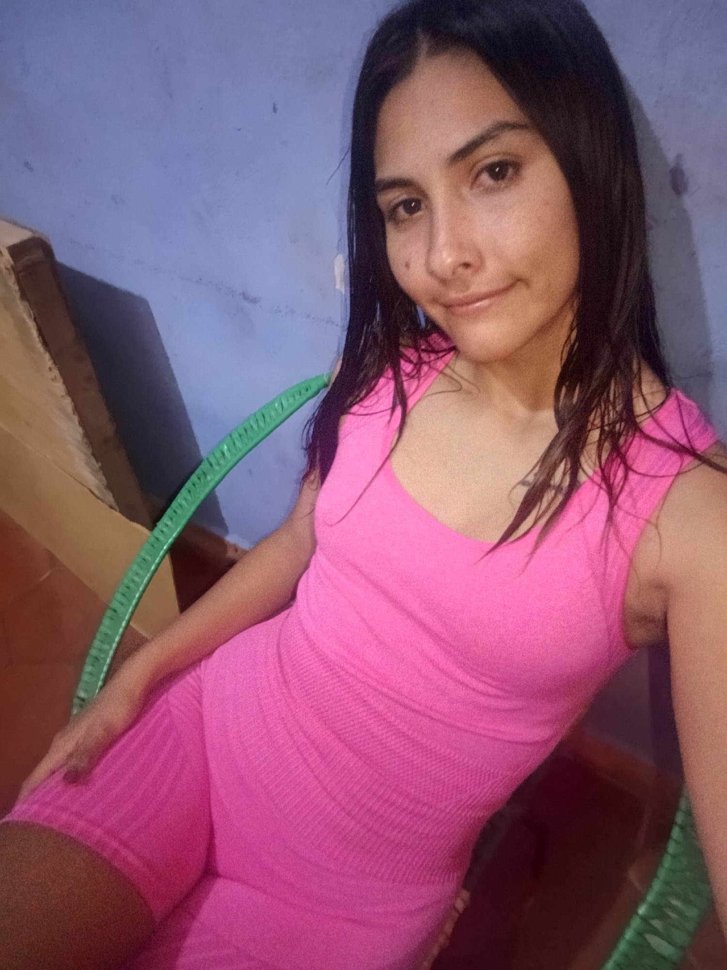 A young woman is captured in a casual, close-up portrait, looking directly at the viewer with a gentle, relaxed smile. She has dark, medium-length hair that appears slightly wet, falling over her shoulders. Her attire consists of a vibrant hot pink, sleeveless top and matching textured shorts, suggesting a comfortable, possibly athletic or leisure wear ensemble. She is seated, with a distinct light green, possibly plastic or wicker, chair visible on the left side of the frame, featuring a ridged design. The background is a textured, faded blue wall, showing some signs of age or wear with darker smudges. A section of a brown structure is visible on the far left. The lighting is soft and even, implying an indoor setting or a shaded outdoor area. The overall mood is calm and natural. The location context for this image is Juan Griego, Venezuela. There is no visible text.