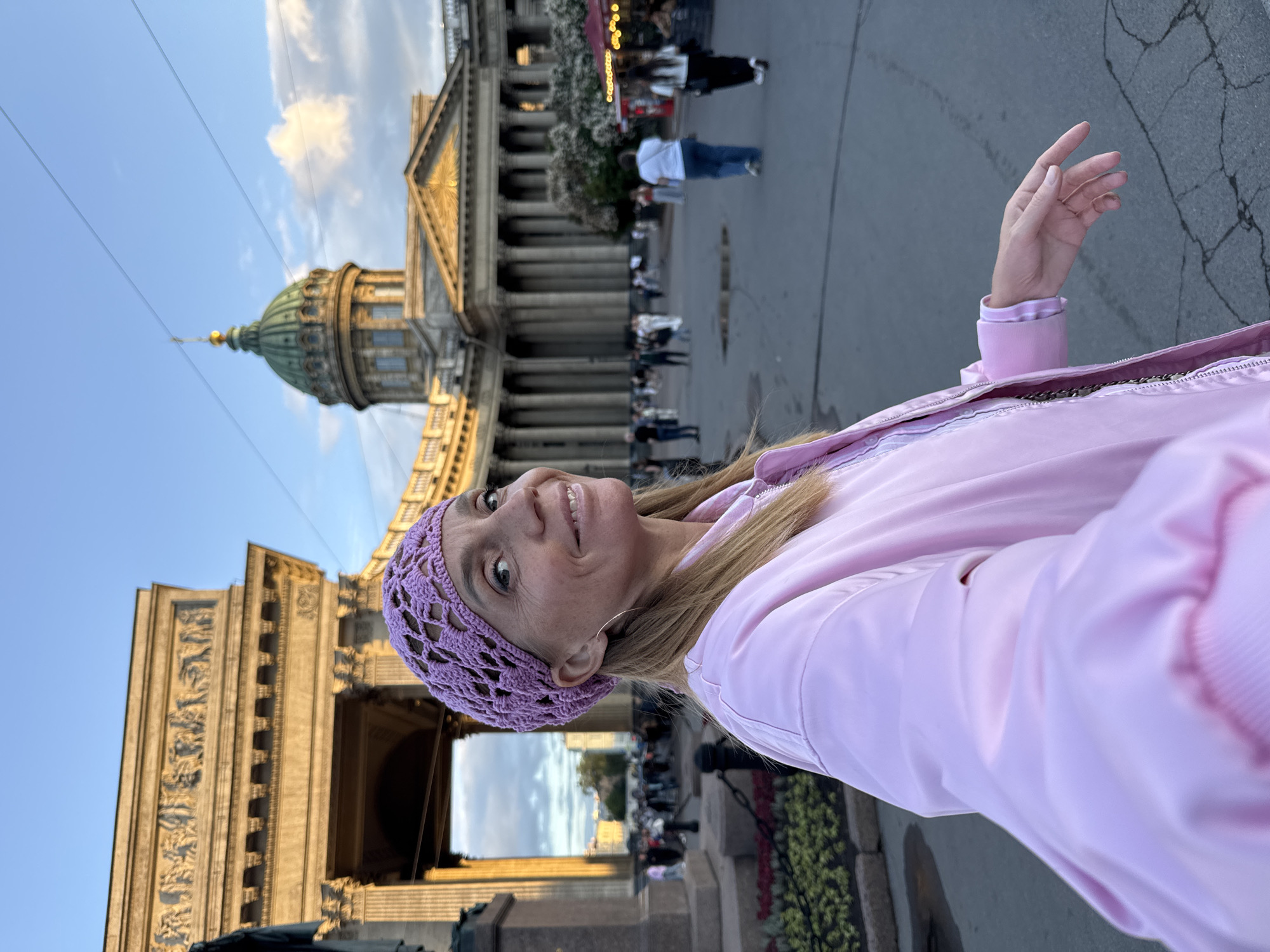 A woman with blonde hair, wearing a pink crocheted hat and a pink bomber jacket, takes a selfie in front of Kazan Cathedral in Saint Petersburg, Russia. The cathedral, with its distinctive colonnade and dome, is visible in the background under a blue sky with scattered clouds. Several people are walking in the square in front of the cathedral. The woman is smiling at the camera, with her right arm extended to take the photo. The lighting suggests it is late afternoon or early evening.