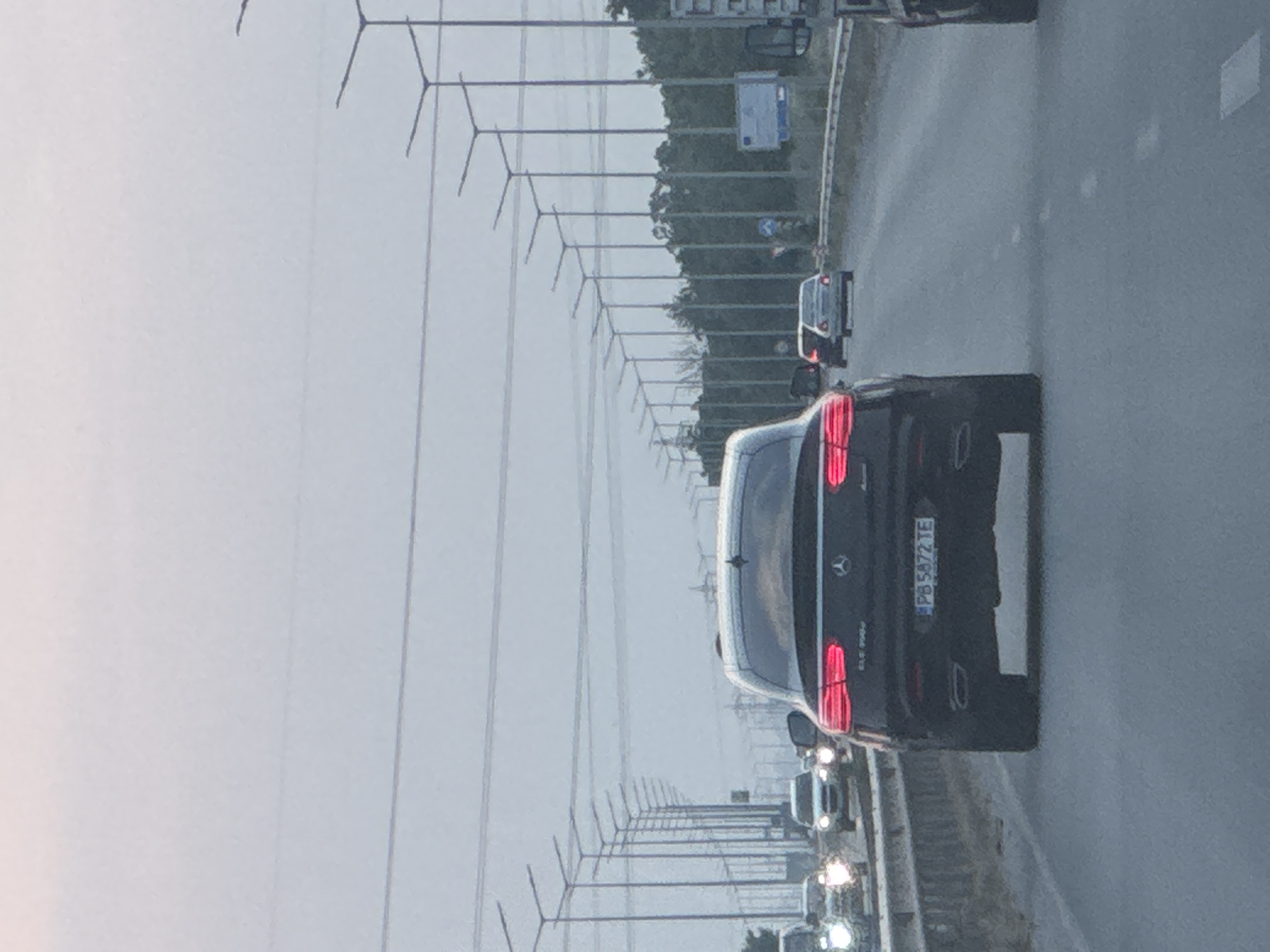 A black Mercedes-Benz GLE 400d Coupe, with illuminated red taillights and dual exhaust tips, is centrally positioned on a multi-lane asphalt highway or major road, heading away from the viewer. Its Bulgarian license plate reads "PB 5872 TE". Ahead, several other passenger vehicles are visible, including sedans and SUVs, along with a white commercial vehicle partially seen on the far right. The road surface is dry, with white dashed lane markings.
Numerous tall, dark grey streetlights with horizontal arms line the right side of the road, extending into the distance. Overhead power lines or communication cables traverse the sky horizontally. A concrete barrier or guardrail runs alongside the road on the right, behind which dense green foliage and trees are visible. Two blue road signs are present on the right shoulder, one square and another rectangular sign bearing the European Union emblem. The sky is uniformly overcast and light grey, indicating diffused lighting conditions. Given the "PB" license plate designation for Plovdiv Province, this scene is located on a highway in or near the city of Asenovgrad, Plovdiv Province, Bulgaria.