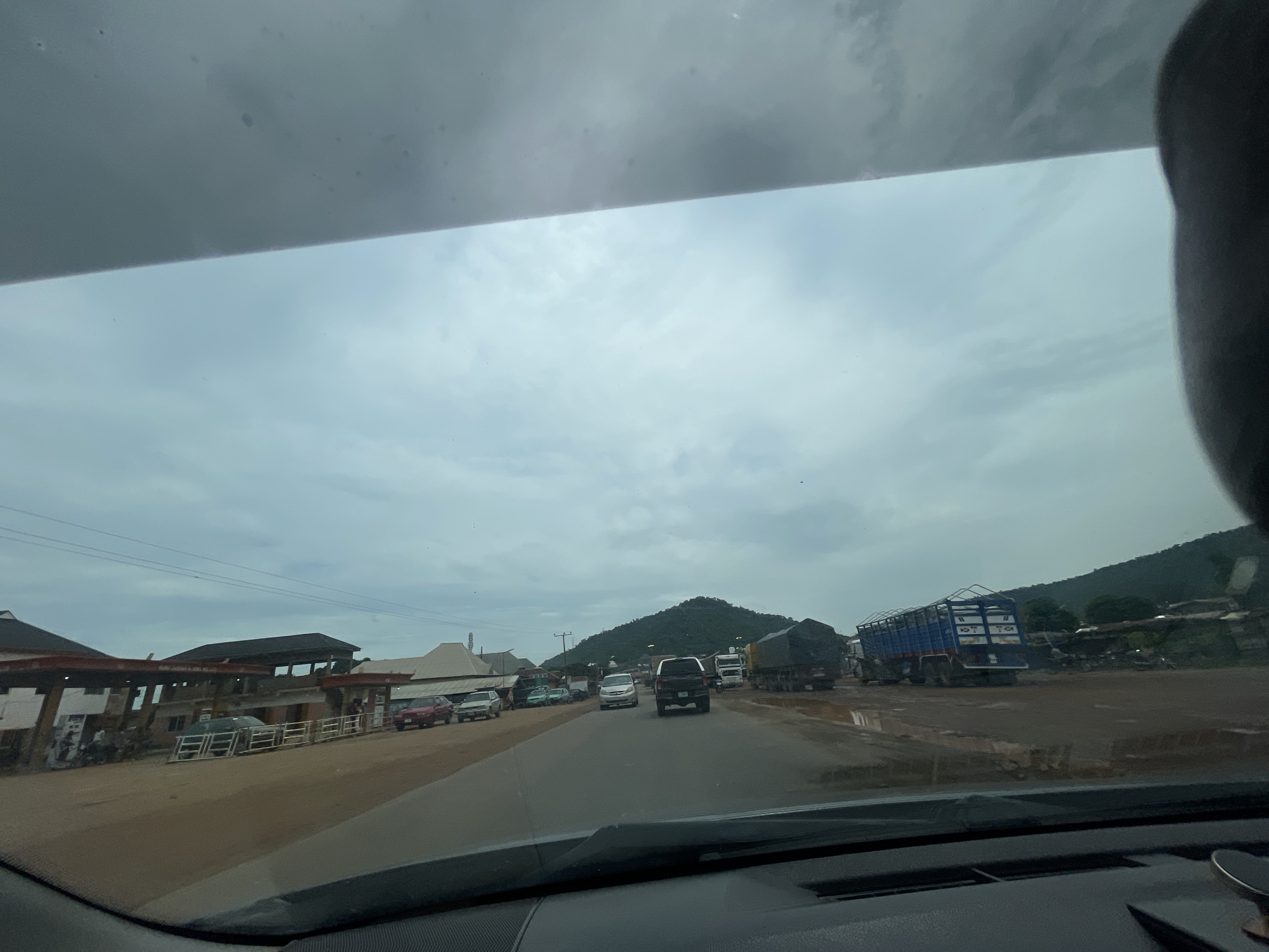 The image depicts a view from inside a vehicle, looking out through the windshield and partly obscured by the car's dashboard. The scene is a road with several vehicles, including cars and trucks, suggesting moderate traffic. On the left side of the road, there are buildings that appear to be commercial establishments, possibly a gas station with multiple pumps visible. Further down the road, a large blue truck with a cage-like trailer is prominent, as are other large trucks, some carrying what appear to be loads covered with tarpaulins. In the background, lush green hills rise against a cloudy, overcast sky. The road surface appears to be asphalt, with some areas showing signs of mud or puddles, indicating recent rain or a damp environment. The overall atmosphere is that of a semi-urban or roadside setting in a tropical or subtropical region, consistent with Koton Karifi, Nigeria. No individuals are clearly discernible in the image.