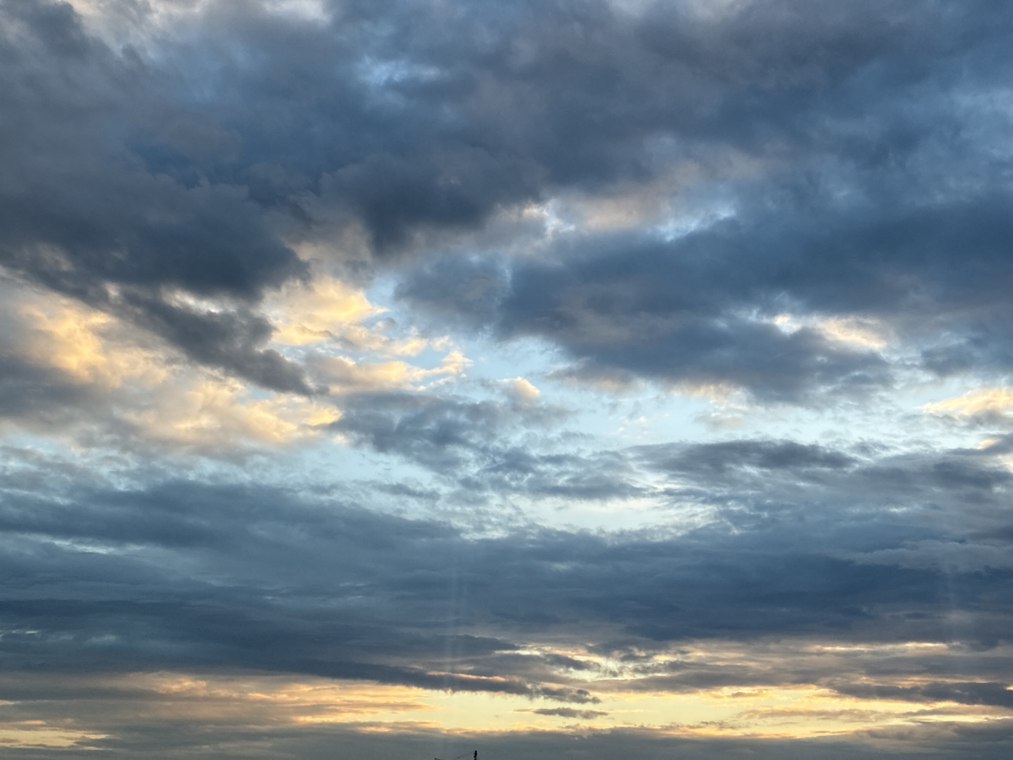 A wide-angle shot captures a dramatic sky filled with a mix of dark, heavy clouds and streaks of golden light, suggesting either sunrise or sunset. The clouds are layered, with lighter, illuminated portions breaking through the darker masses. Subtle shafts of light, possibly crepuscular rays, are visible emanating from behind the clouds in the upper portion of the frame. The scene appears to be an open sky, with no immediate city structures or distinct landmarks visible. The color palette is dominated by blues, grays, and yellows.