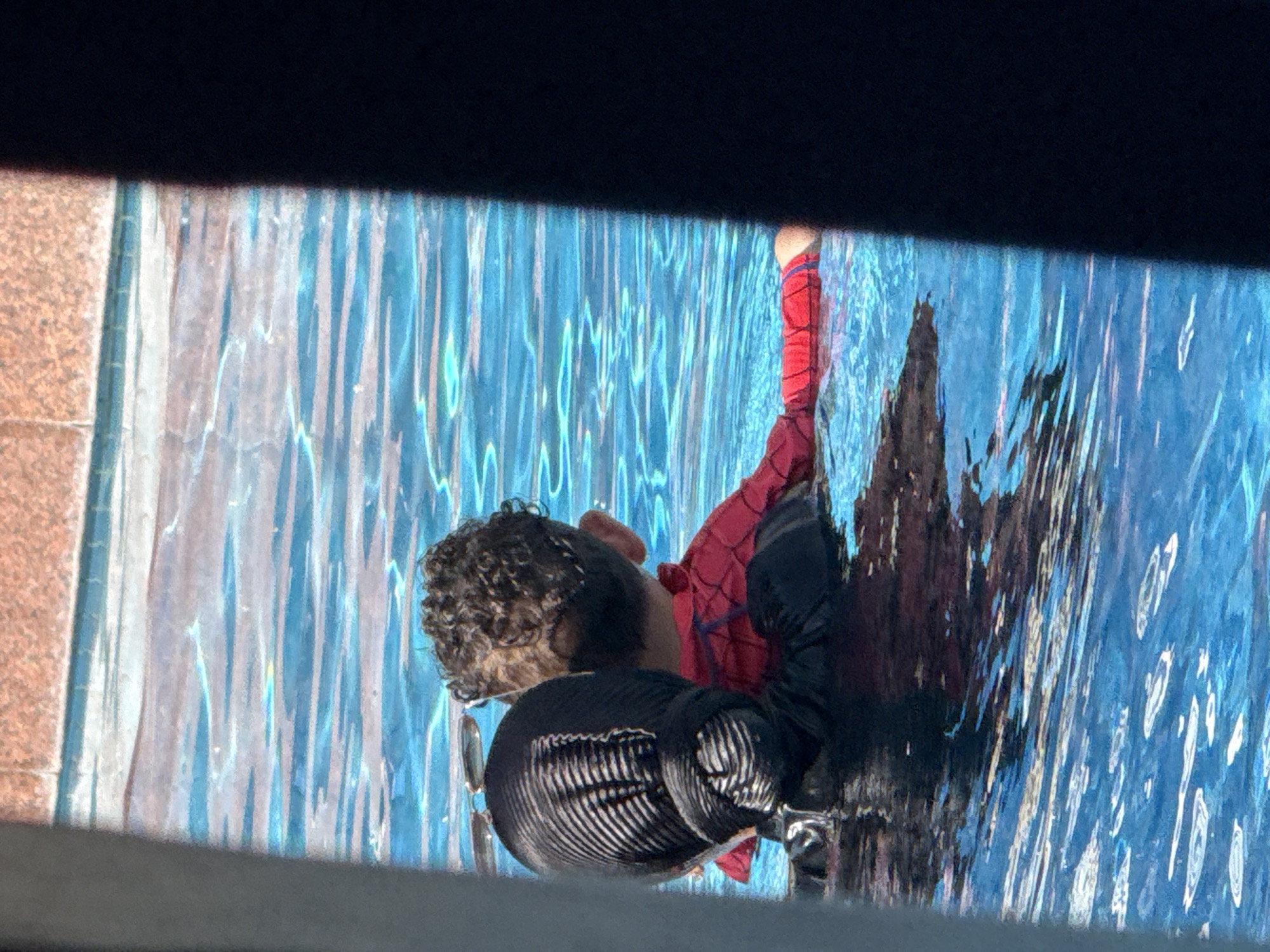 The image shows a person wearing a black headscarf and a red and black swim shirt, likely a child, in a swimming pool. The person is partially submerged in the bright blue water, which reflects sunlight and shows ripples. The edge of the pool, made of reddish-brown tiles, is visible at the top of the frame. The scene appears to be outdoors, possibly in a residential or hotel area, given the tiled pool edge and bright sunlight. There is a dark, vertical shadow or object on the right side of the image, obscuring part of the scene, possibly a wall or a structure. The overall impression is of a leisurely activity, like swimming or playing in a pool. The location is in Dubai, United Arab Emirates.
