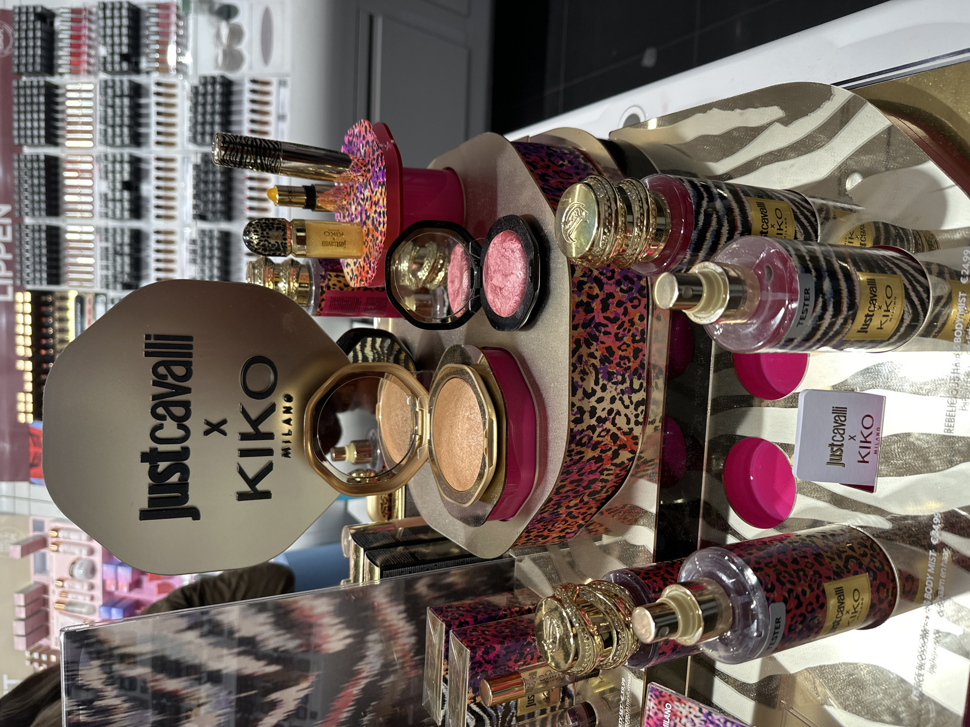 This is a product display of the "Just Cavalli x KIKO Milano" makeup and perfume collaboration. The display features various products, including lipsticks, eye makeup, blush, and body mist. The products are packaged in a distinctive zebra and leopard print design. A display card with the brands "Just Cavalli x KIKO Milano" is visible, along with individual product pricing tags. A tester bottle of body mist can also be seen. The picture was likely taken indoors at a retail location that is selling these products in the city of Amsterdam, Netherlands, given the labels in Dutch.