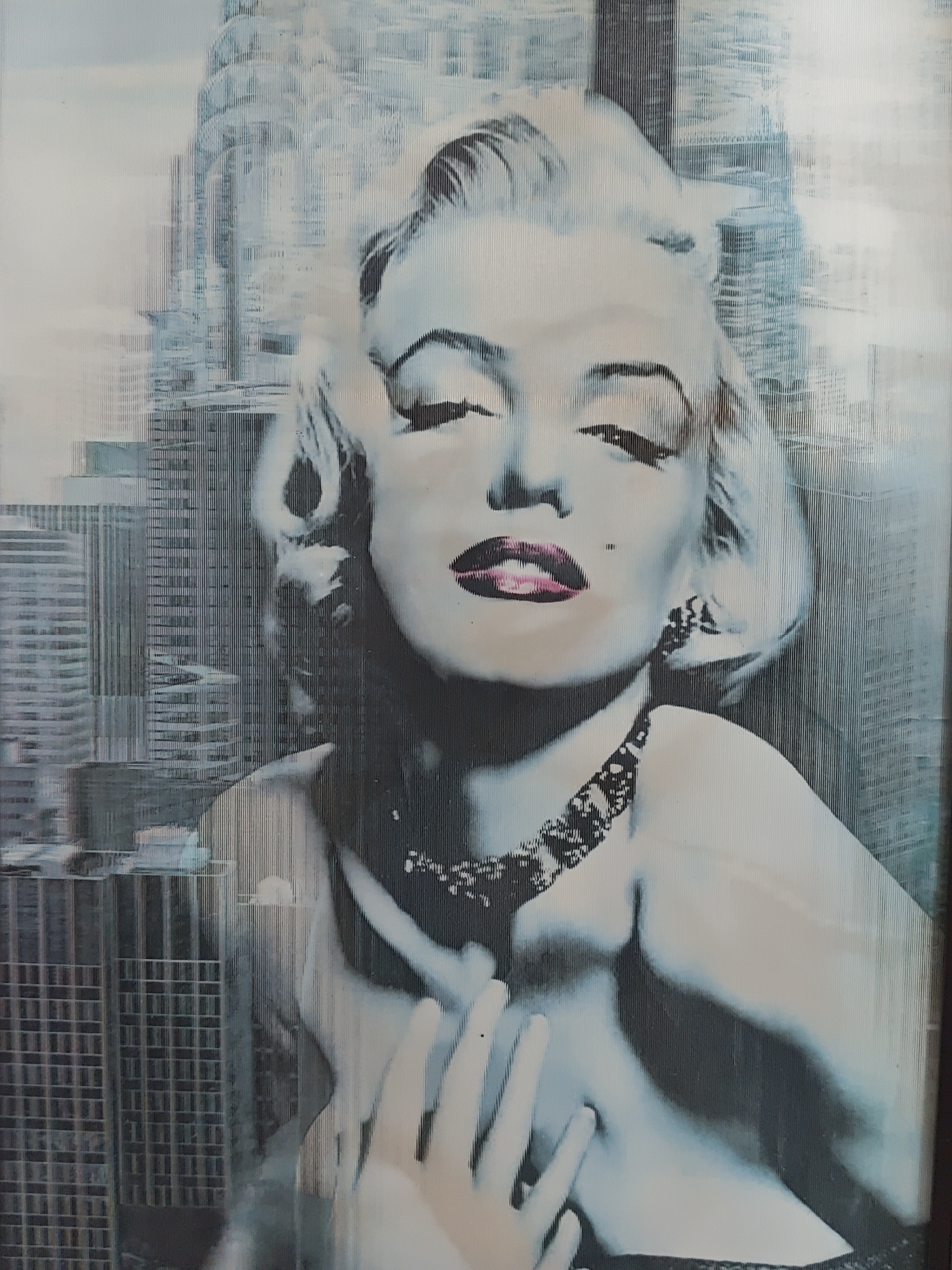 The image is a painting of Marilyn Monroe in a pop art style, superimposed on a cityscape. The painting depicts Monroe with her eyes closed and lips slightly parted, her signature blonde hair styled in soft curls. She is wearing a dark, patterned necklace and a strapless garment. Her left hand is placed gently on her chest. The background is a stylized representation of skyscrapers, rendered in shades of blue and grey, with vertical lines that create a sense of texture and depth. The overall impression is one of glamour and urban energy, with the iconic figure of Marilyn Monroe set against a backdrop of a modern metropolis. The painting does not depict any specific location in Saint Petersburg, Russia.