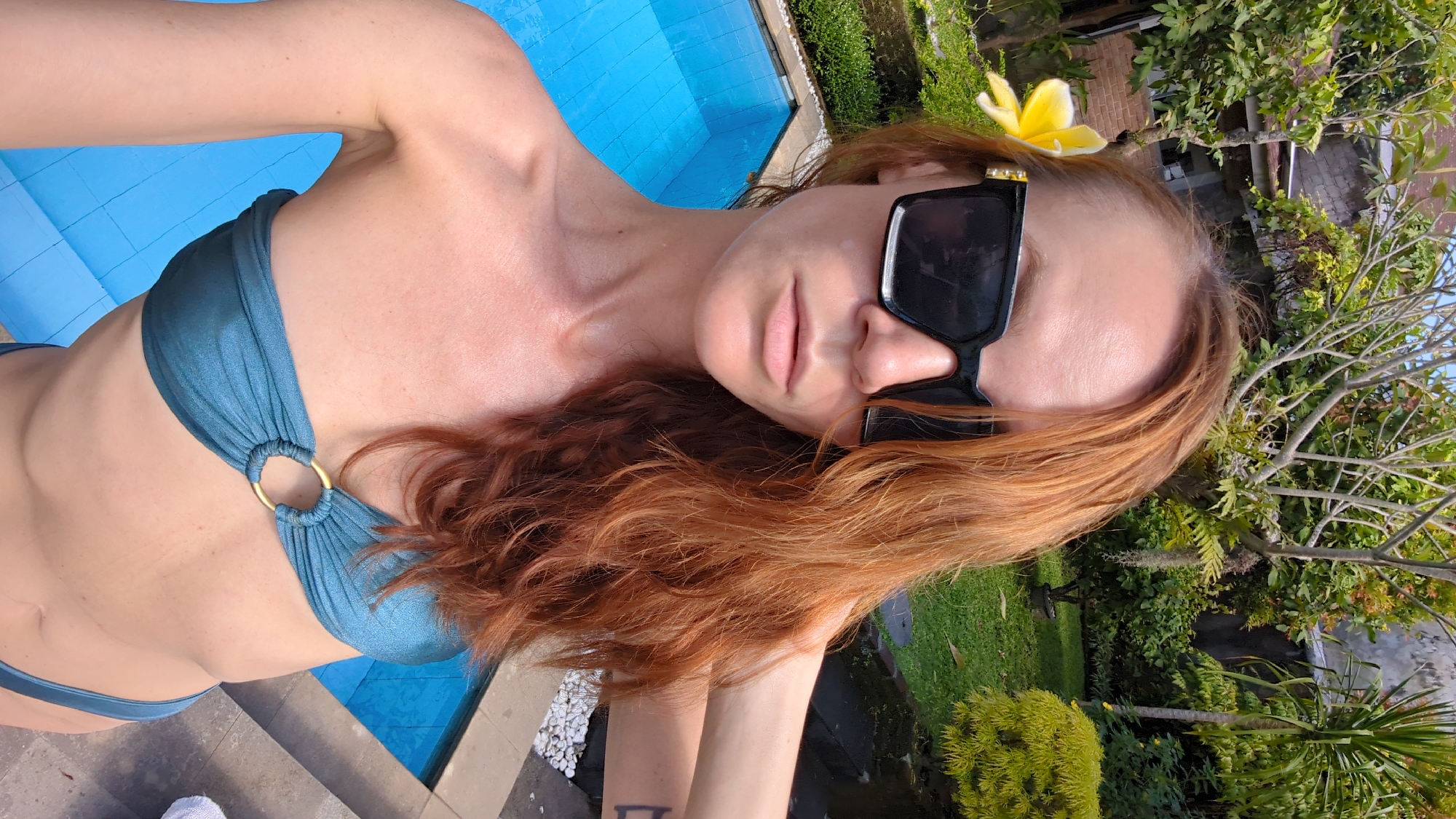 The image captures a close-up, high-angle selfie of a woman relaxing by a swimming pool in a tropical outdoor setting. The woman, with long, wavy reddish-brown hair, is wearing oversized black sunglasses and a teal or turquoise bikini top with a prominent gold O-ring detail at the center. A bright yellow frangipani flower is tucked into her hair above her right ear, enhancing the tropical theme.
Her skin, visible on her arms, neck, and torso, appears slightly glistening or flushed, indicative of sun exposure. She is positioned next to a vibrant blue swimming pool, part of which is covered by what looks like a blue mat or float. Concrete steps are visible leading into the pool near her body.
The background features lush, dense green tropical foliage, including trees, shrubs, and grassy areas, suggesting a garden or resort environment. White decorative pebbles are visible along the edge of the pool. The scene is brightly lit by natural sunlight, indicating a clear, sunny day, likely during mid-morning or late afternoon given the soft shadows. The overall mood is relaxed and leisurely, consistent with a vacation in a warm, exotic location like North Kuta, Indonesia. No discernible text is visible within the image.